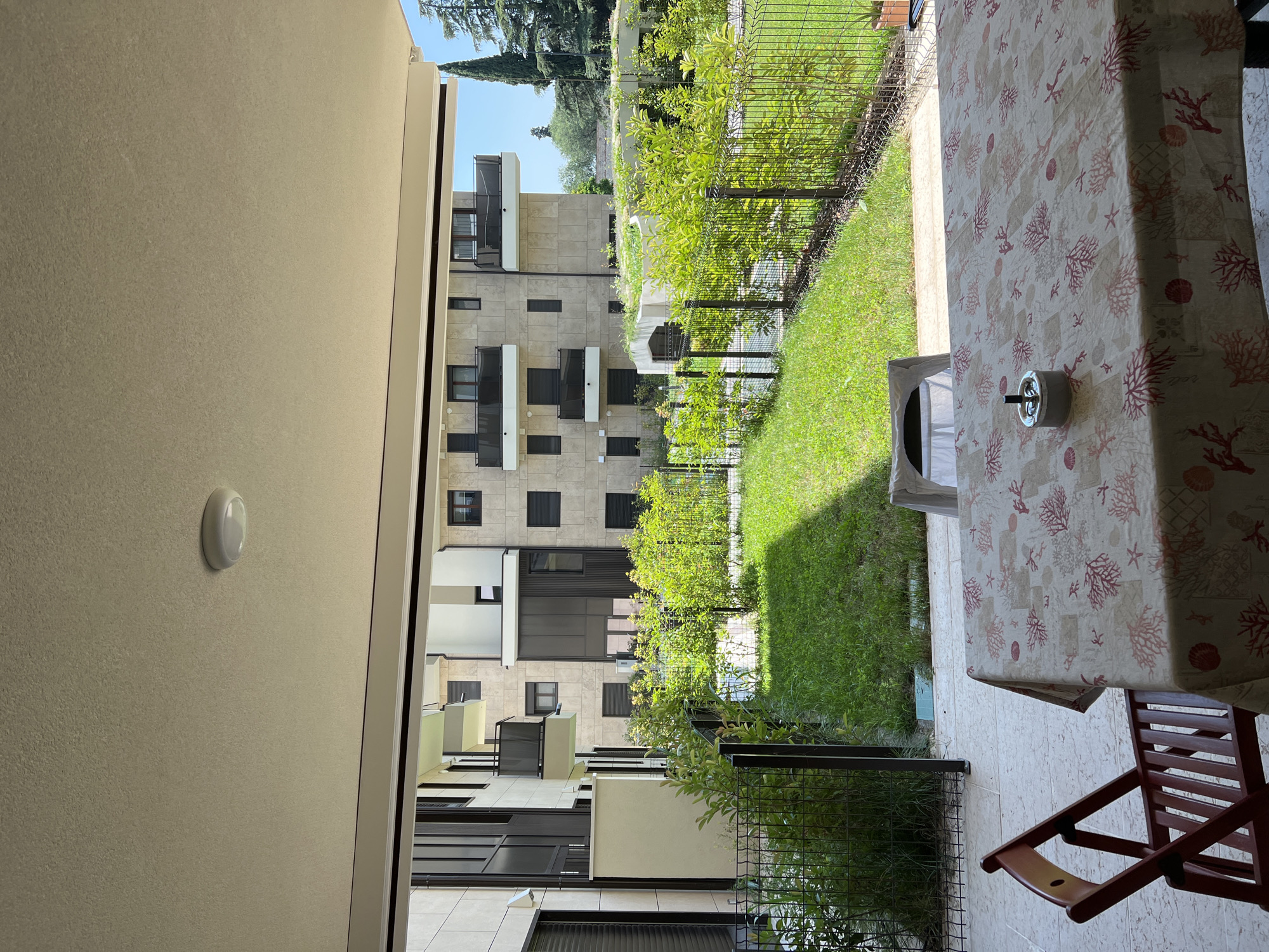 The image shows a view from a covered patio looking out onto a small, manicured lawn and a modern apartment building. A table covered with a coral-patterned tablecloth is in the foreground, with a folding wooden chair to its left. An ashtray sits on the table. The building in the background appears to be made of light-colored stone and features multiple balconies and dark-tinted windows. A lush green hedge runs along the edge of the lawn, separated from the patio by a black metal fence. The sky is bright blue with a few scattered clouds, suggesting it is a sunny day. The presence of the building and its modern design suggests an urban or suburban setting. Given the context of the city of Verona, Italy, this scene likely depicts a residential area, possibly an apartment complex, with a communal garden space.