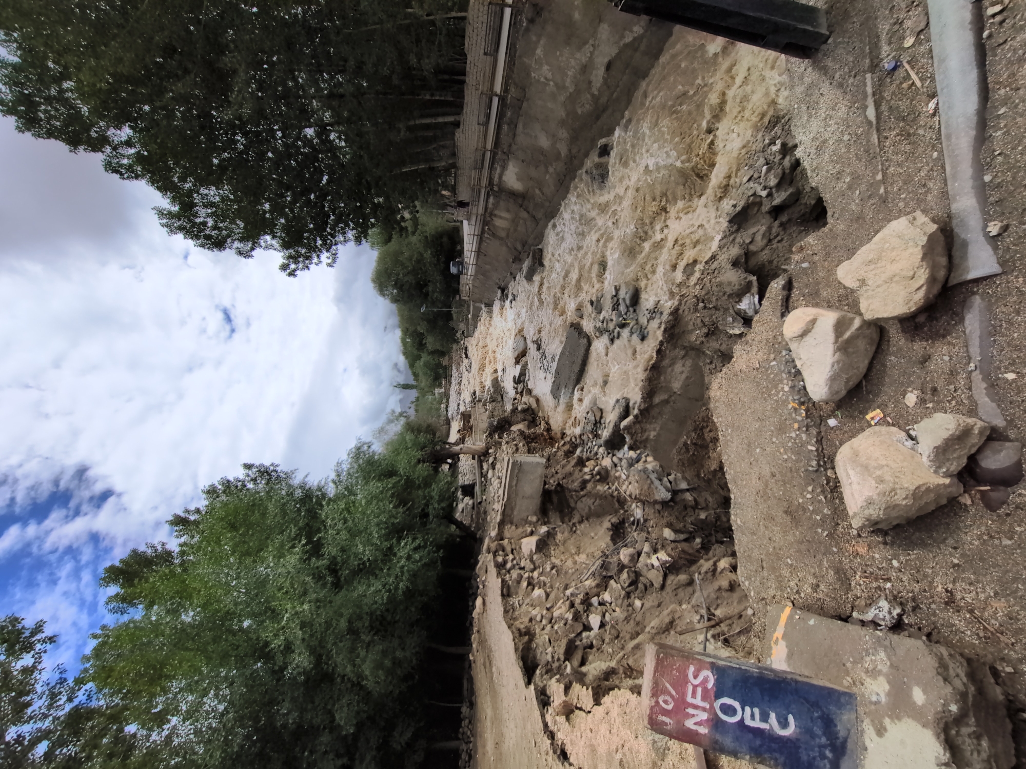 The image depicts the aftermath of a flood or landslide in Leh, India. A muddy, fast-flowing river has eroded a significant portion of the land, creating a deep chasm. On the left side of the image, a road or pathway has been severely damaged and collapsed into the chasm. A concrete structure, possibly part of a bridge or retaining wall, is partially visible in the eroded area.
A marker with the text "JO/NFS/OFC" in white on a blue and red background stands precariously on the edge of the collapsed ground, indicating a boundary or signpost. The riverbank on the right side of the image appears to be reinforced with a concrete channel or wall, and a walkway or road runs alongside it, with two people visible in the distance walking along it. Trees line the far side of the river and the distant landscape, under a cloudy sky. The scene conveys destruction and the power of natural forces.