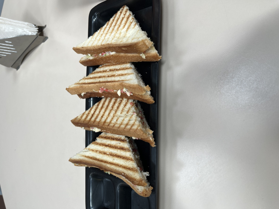 Four grilled cheese sandwiches are arranged in a row on a black tray, which is sitting on a beige table. The sandwiches are cut in half and the filling is visible. They are arranged from left to right, with the first sandwich in the row being slightly offset from the rest of the sandwiches. The scene is located in Sahibzada Ajit Singh Nagar, India. The coordinates indicate that the scene is likely indoors. The sandwiches are the only subjects in the image. There is no evidence of any people or interactions. The lighting is bright and even, with a slight reflection on the surface of the table. The background is a simple white table with a light grey countertop. The environment appears clean and uncluttered. There is a small metallic object in the upper right corner of the image, which could be a napkin holder.