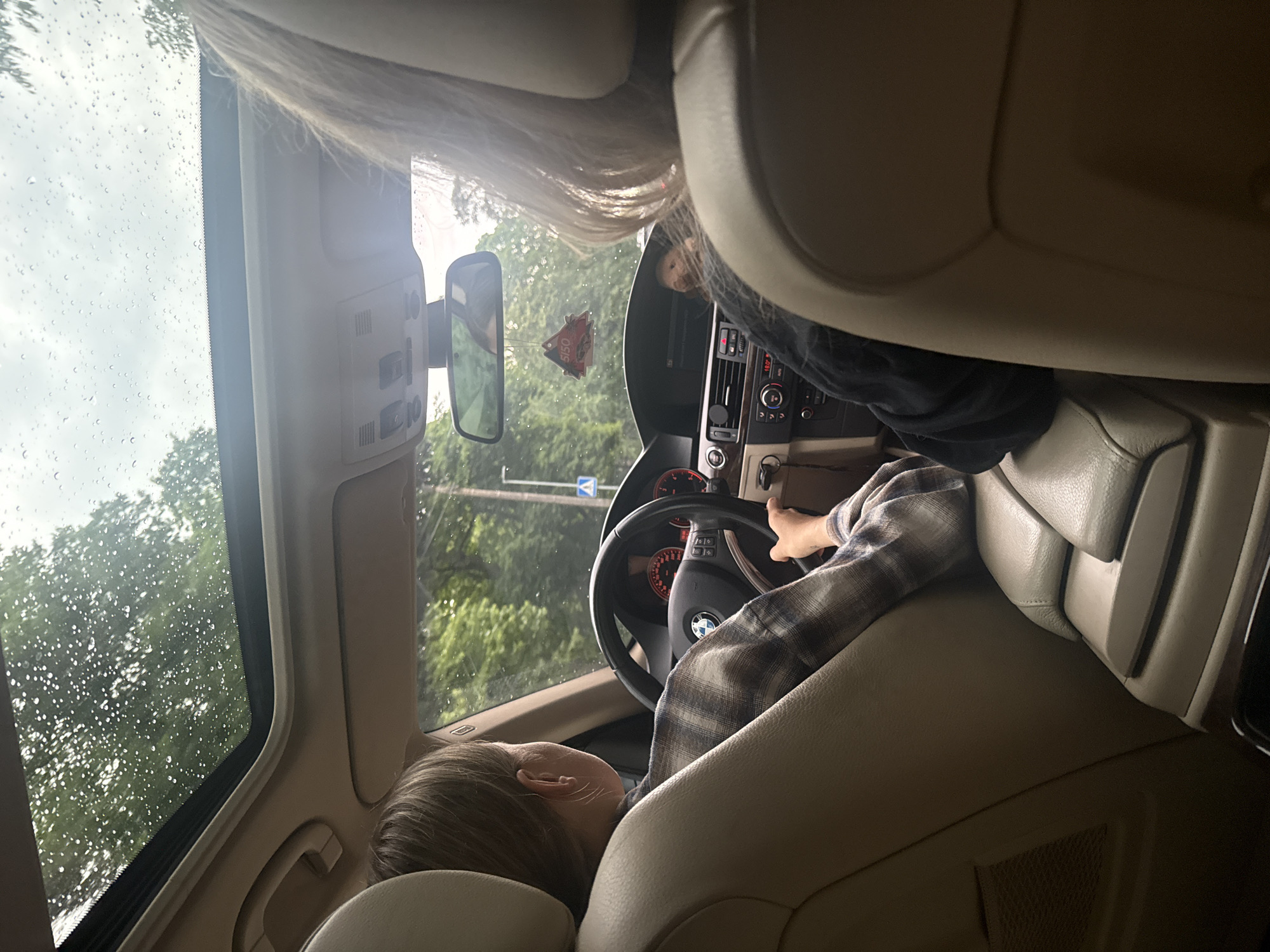 In the image, two individuals are seated inside a car. The driver, a woman with light brown hair, is positioned in the front seat on the left, her hands on the steering wheel. She is wearing a plaid shirt. To her right, another person, with blonde hair, is visible from the back, facing forward. The car's interior features a sunroof with raindrops on it, suggesting inclement weather. The dashboard, steering wheel, and car seats are clearly visible. Outside the car, trees and a road can be seen through the windows and sunroof, indicating they are in transit. There's a car air freshener hanging from the rearview mirror. The overall scene captures a moment during a car ride, possibly on a rainy day.