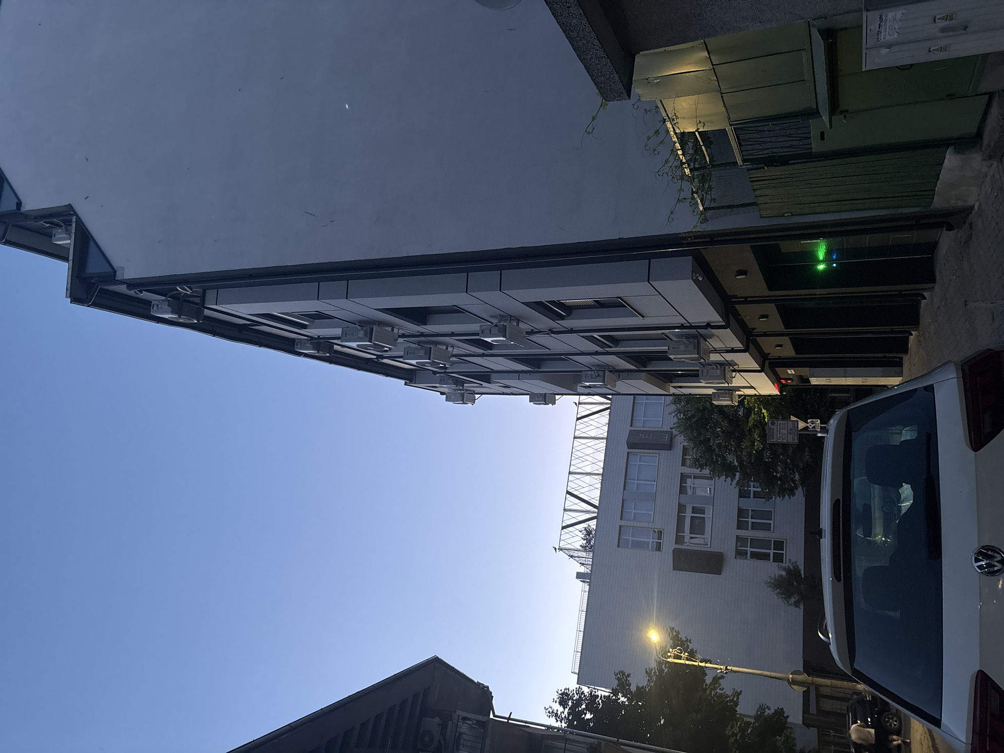 The image depicts a street scene in Burgas, Bulgaria, likely during dusk or early evening, as indicated by the blue sky and artificial lighting. In the foreground, the rear of a white Volkswagen SUV is visible. To the right, a multi-story building with a light-colored facade dominates the frame, featuring numerous air conditioning units mounted on its exterior. The building's facade is largely smooth, with some darker paneling or window frames.
Further into the scene, to the left, another light-colored building with multiple windows is visible, partially obscured by trees. A streetlamp with a warm yellow glow illuminates the area near this building. Across the street, another building is partially visible, with a darker, angled roofline.
The ground appears to be paved, suggesting a pedestrian walkway or sidewalk. In the mid-ground, a narrow alley or pathway runs between buildings. There is a green metal fence and some doors visible in this area, with a bright green light emanating from one of the building's entrances.
Two individuals are casually walking on the left side of the street in the mid-ground. One is wearing shorts and a t-shirt, and the other is in the distance. The overall impression is an urban environment, typical of a residential or commercial area in a city.