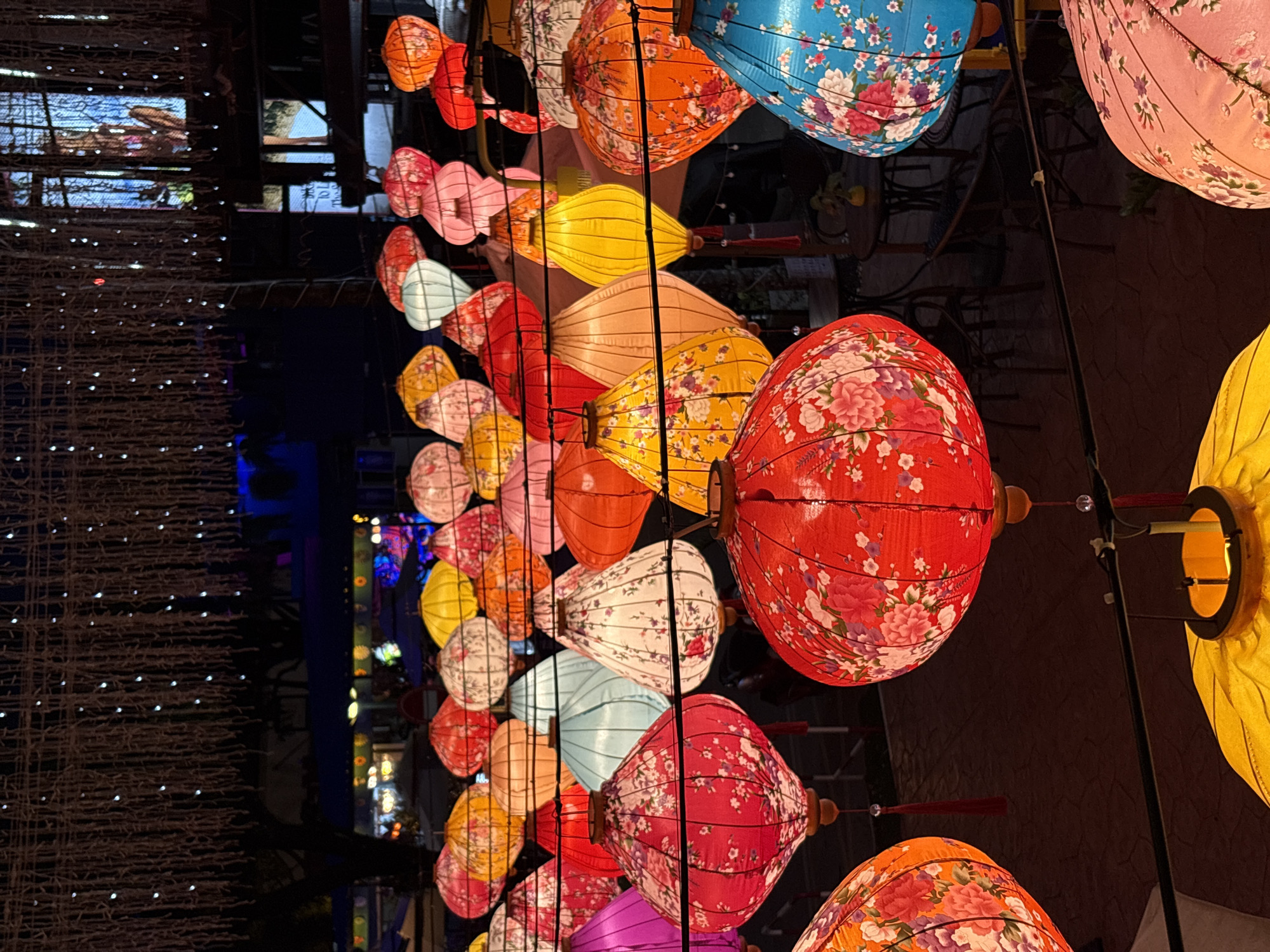 The image depicts an outdoor nighttime scene in Choeng Thale, Thailand, featuring a vibrant display of numerous hanging lanterns. The lanterns are of various sizes, shapes, and colors, predominantly red, pink, yellow, orange, and blue, many adorned with floral patterns. They are illuminated from within, casting a warm glow. In the background, there are blurred lights and structures, suggesting a street or market area with some trees and decorative string lights overhead. The ground appears to be paved. No people are clearly visible in the frame. The overall atmosphere is festive and decorative.
