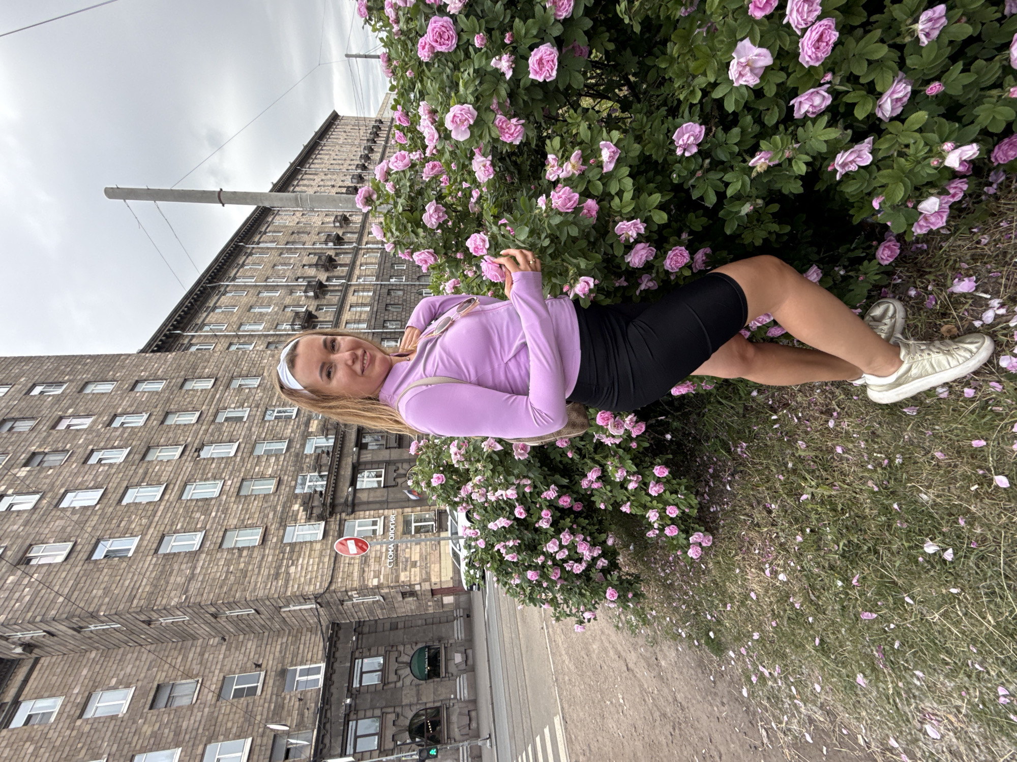 A blonde woman with a headband stands in front of a bush of pink roses on a cloudy day. She is wearing a lavender athletic jacket and black shorts. Behind her is a large, stone building and a street with a pedestrian crossing. The ground is covered in fallen rose petals.