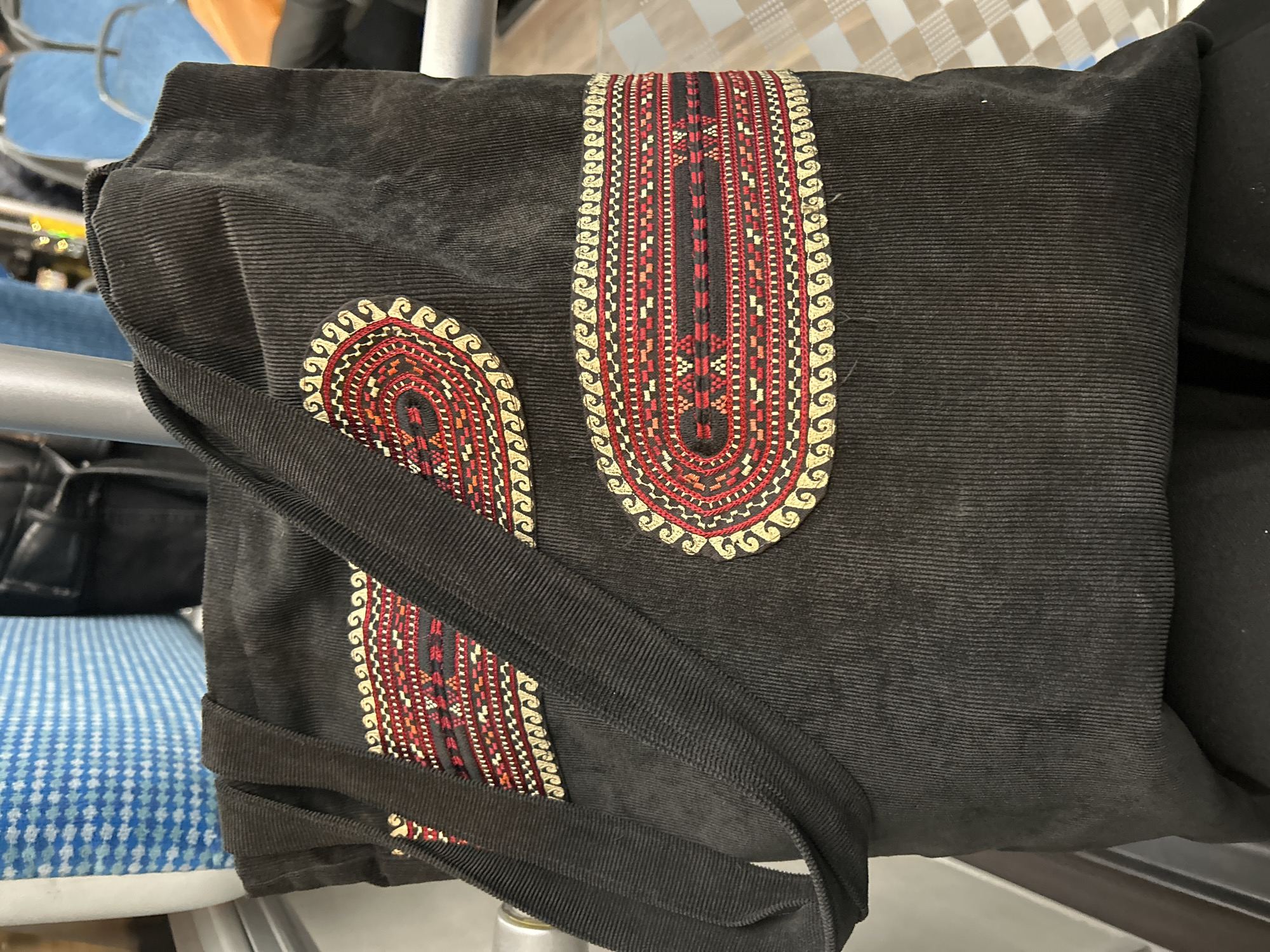 A dark, corduroy tote bag with embroidered accents sits on a person's lap in a public transportation setting. The bag features two horizontal embroidered bands with intricate red, yellow, and black geometric patterns. The background shows blue and white patterned upholstery and metal railings, suggesting the interior of a bus or train. The scene is indoors, and the lighting appears to be artificial. The person's lower body is visible, indicating they are seated.
