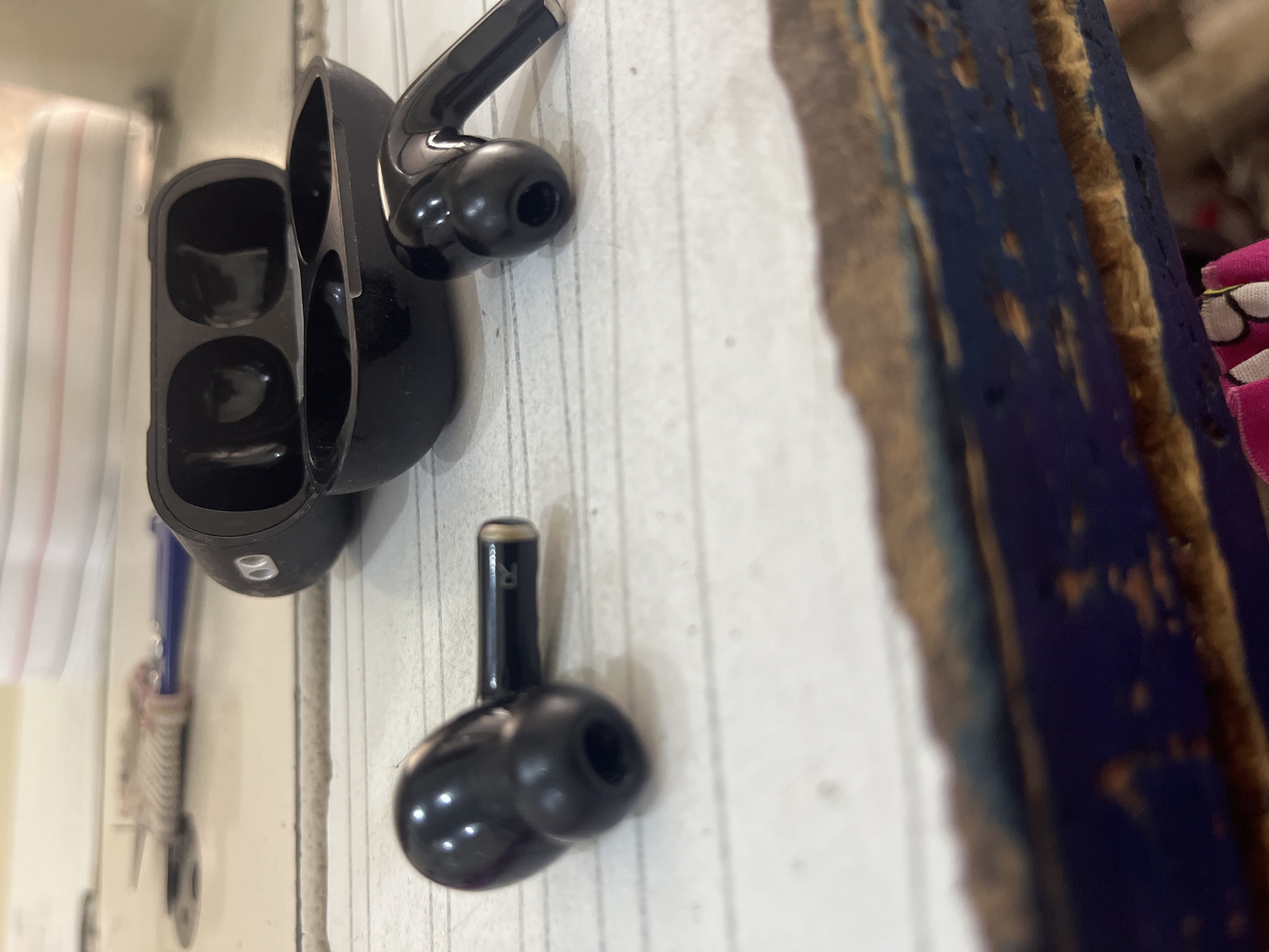 The image shows a pair of black wireless earbuds and their charging case on a white, lined surface. One earbud is positioned to the left, and the other is to the right, close to the open charging case. The charging case is black and has two indentations for the earbuds. The surface appears to be a table with a distressed, wooden edge in the foreground. In the background, out of focus, there is a white surface with thin red and blue stripes, and a blue tool with a brush-like attachment.