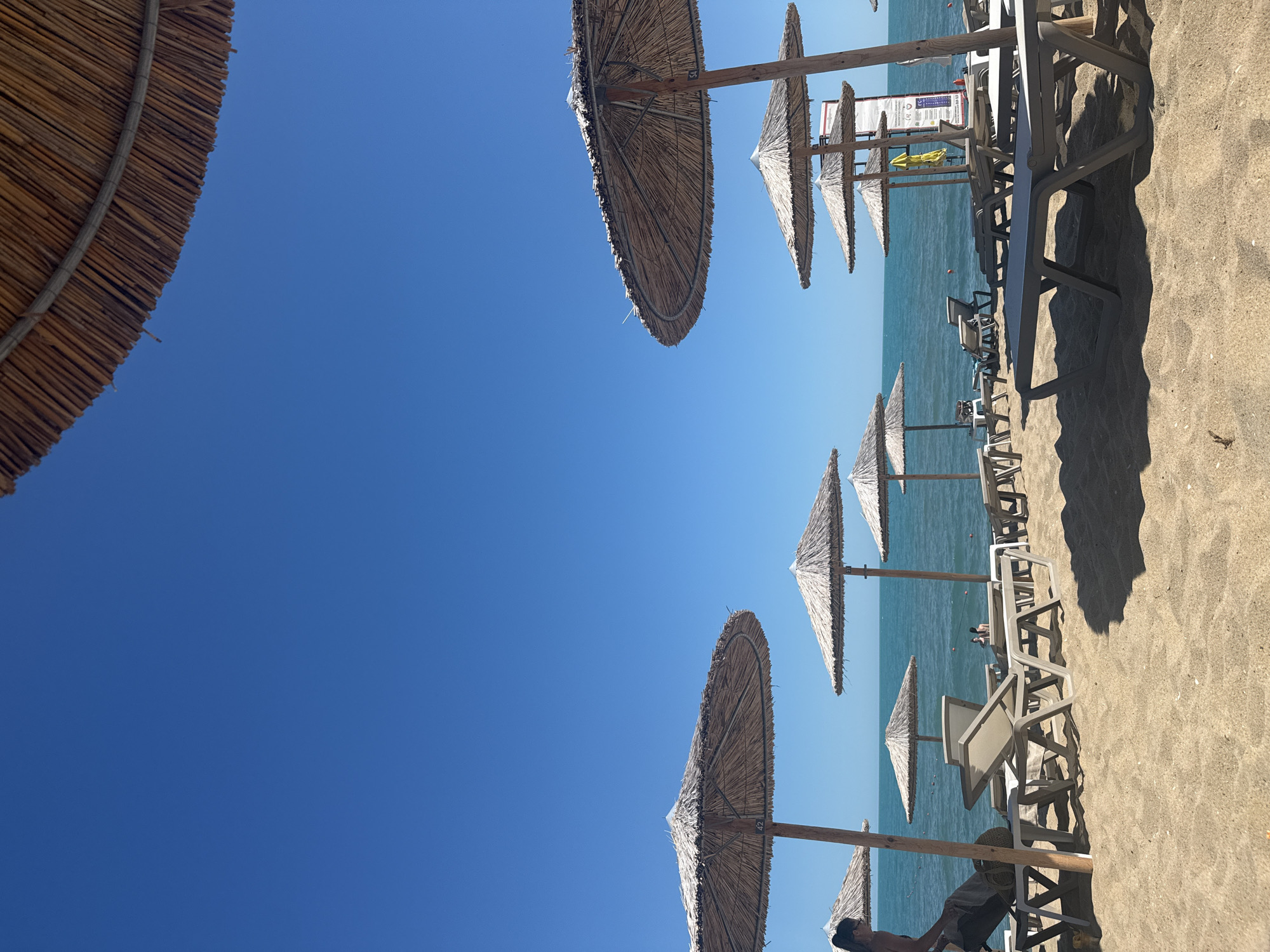This image depicts a sun-drenched beach scene with rows of straw umbrellas and lounge chairs set up on a sandy shore. The turquoise water of the Black Sea stretches out to the horizon under a clear blue sky. Several beachgoers are visible, with one person in the foreground on the left, appearing to be preparing lounge chairs. Others are seen in the water, swimming or wading. A lifeguard stand with a sign is positioned further down the beach. The scene suggests a peaceful and relaxing day at a resort or public beach in Varna, Bulgaria.