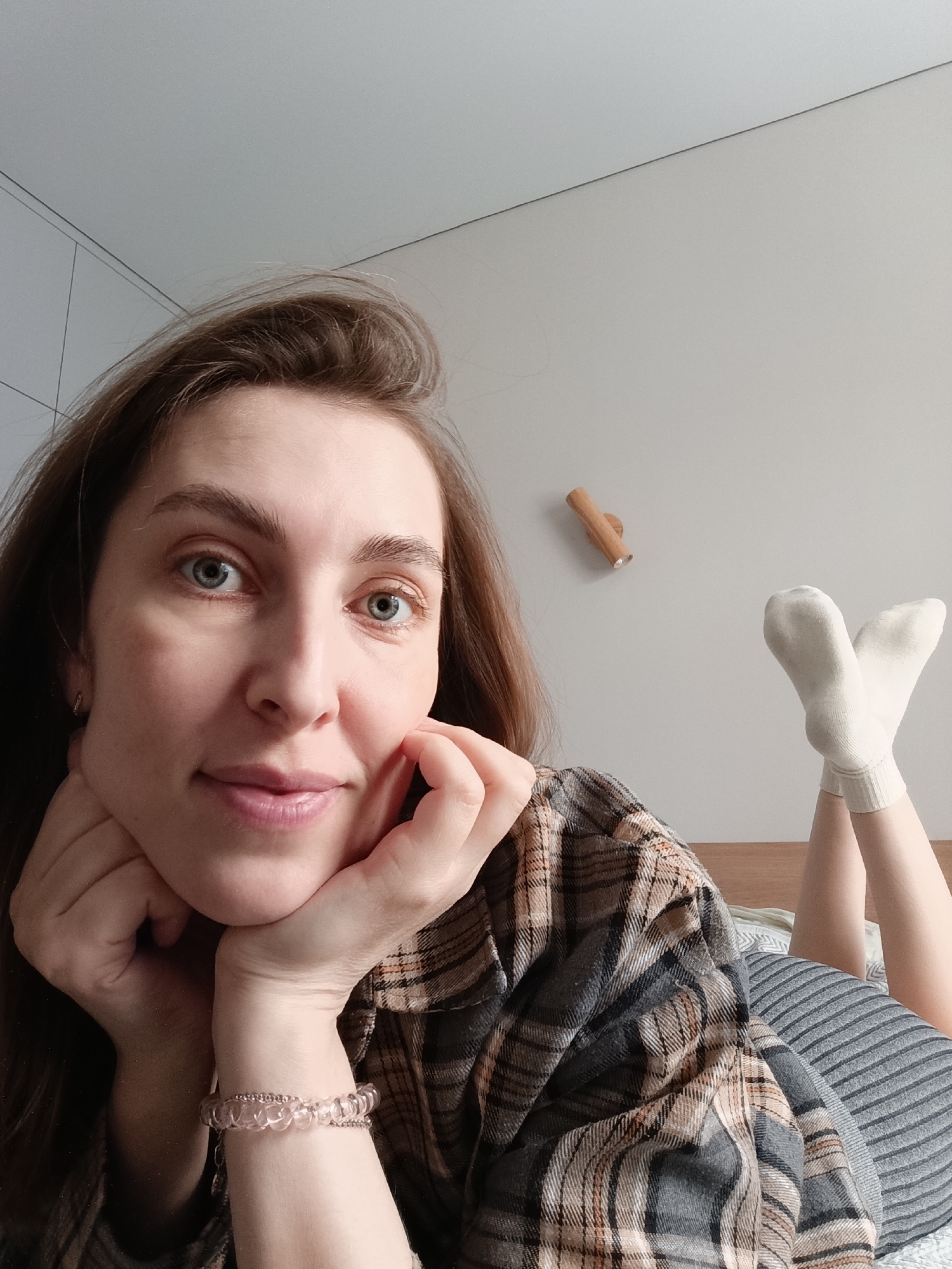A young woman with long brown hair and blue eyes lies on her back in bed, resting her head in her hands. She is wearing a plaid shirt and striped clothing. White socks are visible on her feet, which are raised in the air. The room has a neutral wall color and minimal decoration, with a wooden knob-like object mounted on the wall behind her. The image was taken indoors, likely in a residential setting. Location is Chaikovsky, Russia.