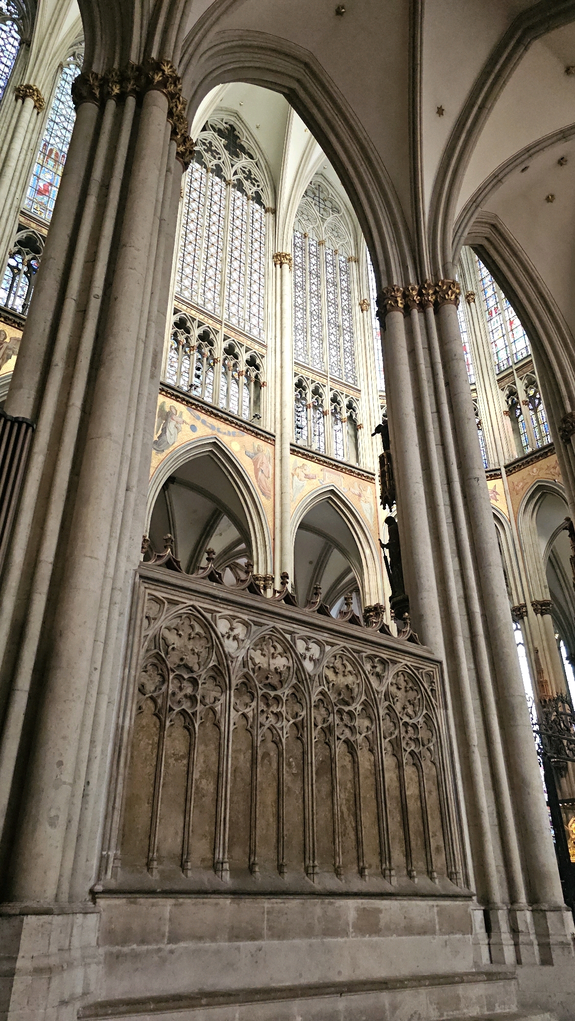 The image captures the interior of a grand Gothic cathedral, likely Cologne Cathedral, Germany. Large, fluted stone columns with ornate gold capitals frame the view. Behind the columns, towering arched windows with intricate tracery and stained glass are visible, letting in diffused light. Below the windows, decorative friezes and arches are present. A prominent feature in the foreground is a stone screen with elaborate Gothic carvings, creating a sense of depth and architectural detail. The overall impression is one of immense scale, historical grandeur, and intricate craftsmanship characteristic of Gothic architecture.