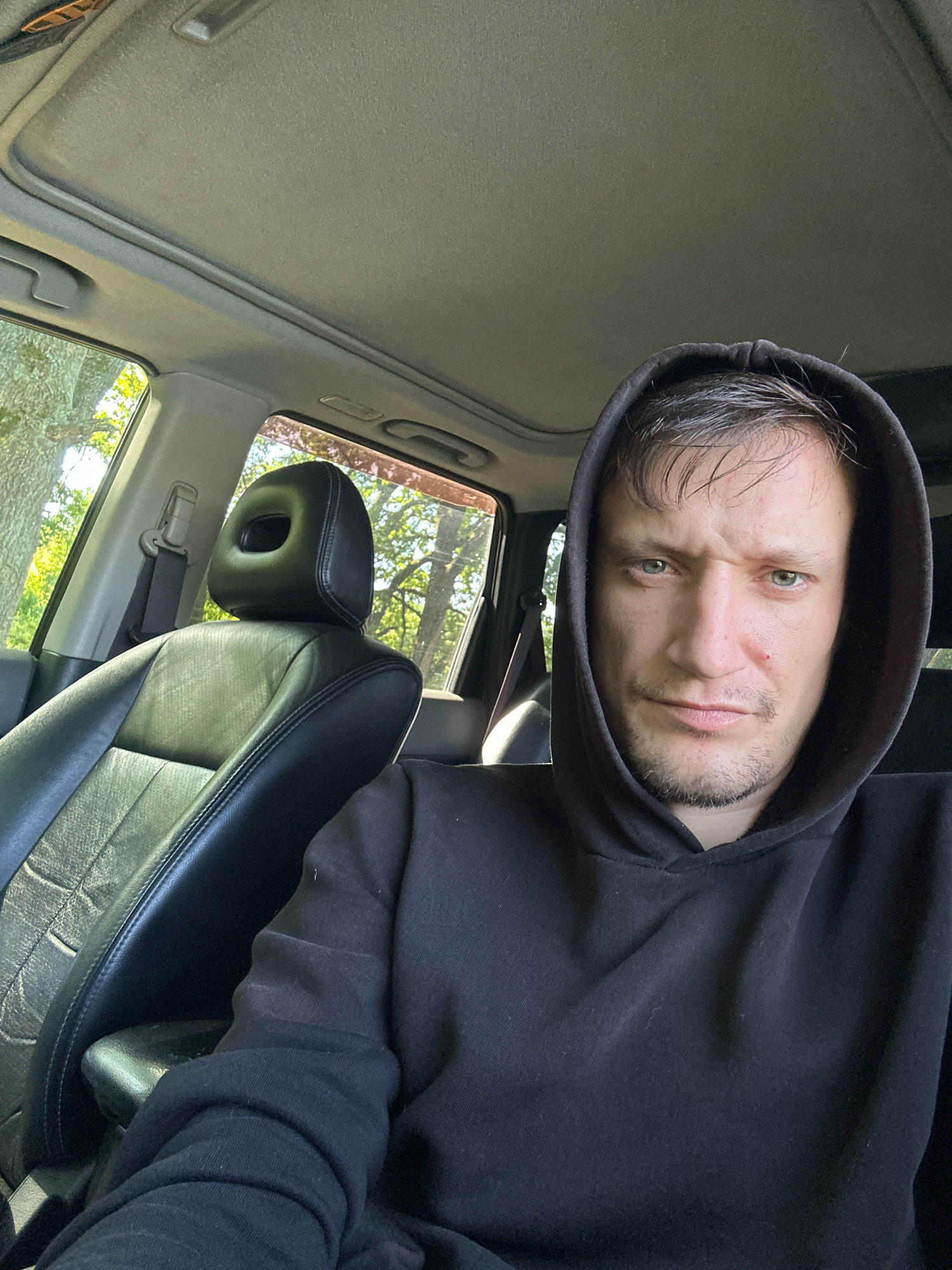 A selfie is taken from inside a car, capturing a man with short brown hair and light green eyes. He is wearing a black hoodie and has a slight stubble. The man's face is turned towards the camera with a neutral expression. To the left of the man, a black leather car seat is visible, with a portion of the headrest also in frame. The car's interior appears to be illuminated by sunlight, suggesting it is daytime. Trees and foliage are visible through the car windows, indicating an outdoor setting, possibly in a rural or suburban area. The provided context states the location is Shatava, Ukraine.