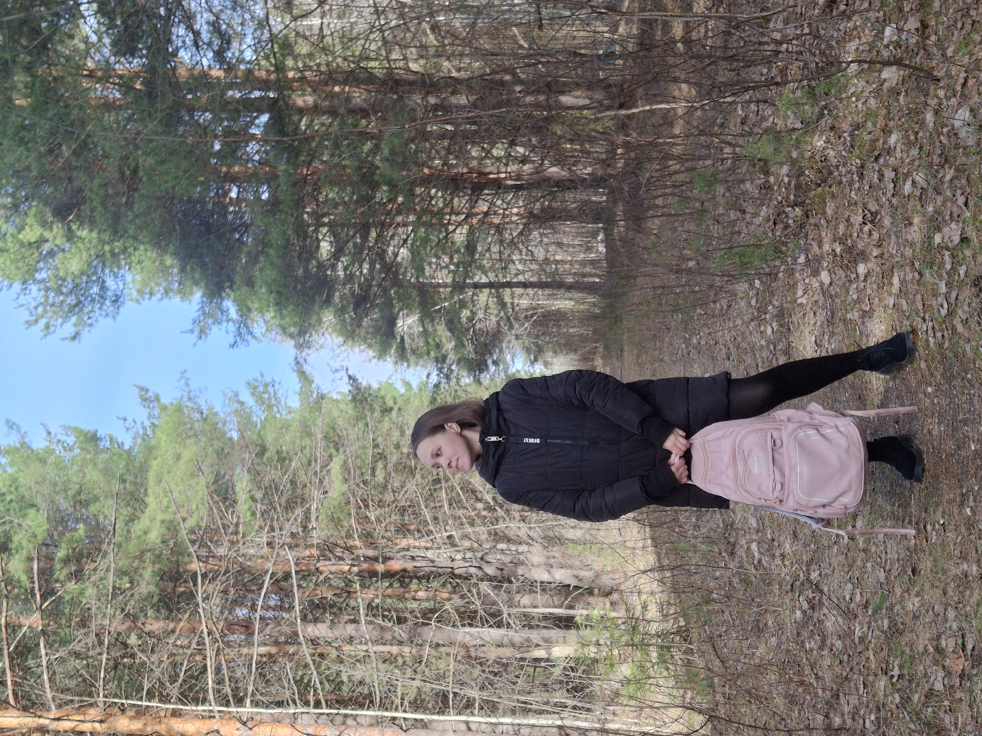 A young woman stands on a narrow path through a forest, likely in Chaikovsky, Russia. The ground is covered with fallen leaves and small branches. The woman is wearing a black coat, dark leggings, and black shoes. She holds a pink backpack. The forest surrounding her is dense with bare trees and evergreens, and the sky is visible through the branches.
