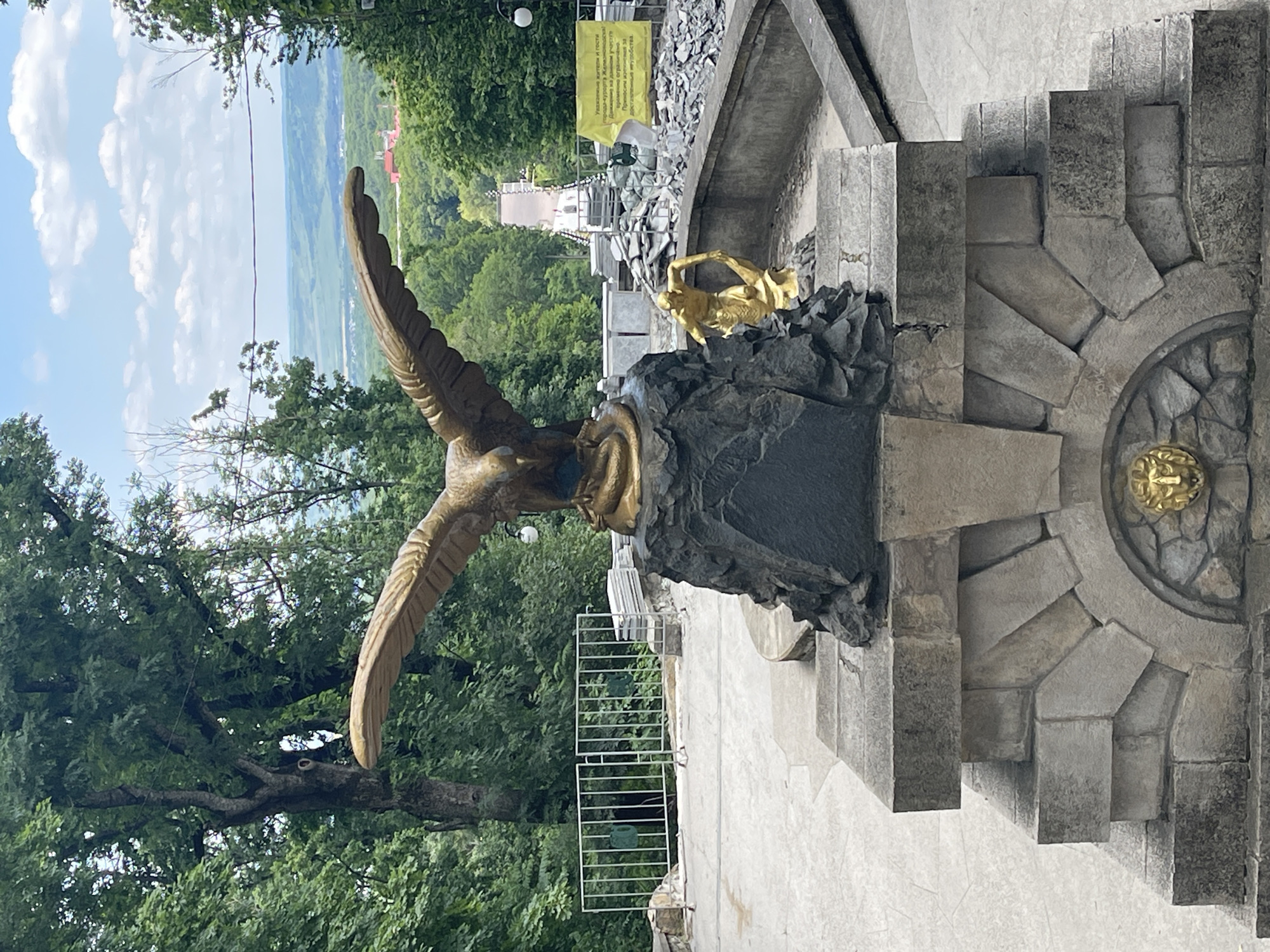 This media file captures a scene featuring a monumental bronze eagle statue with outstretched wings, positioned in an outdoor setting. The statue is mounted on a dark, rocky base and is adorned with golden accents, particularly on a smaller golden figure at its base. The immediate surroundings consist of stone structures, including carved steps and a circular stone element with a golden lion's head.
In the background, a lush green landscape unfolds, with rolling hills and trees under a bright, cloudy sky, suggesting a daytime setting with fair weather. A white railing is visible, hinting at a viewing platform or walkway. Beyond the natural scenery, there are glimpses of buildings, some with red roofs, and a distant tower or structure.
A yellow sign with text is prominently displayed behind the statue, providing context about temporary restrictions. The visible text, in Russian, reads: "Dear residents and guests of the resort town of Zheleznovodsk, passage in this area is temporarily limited. We apologize for the inconvenience." This indicates the location is Zheleznovodsk, Russia, and suggests the area may be undergoing some form of construction or maintenance. The overall impression is a scenic, monument-rich location with a hint of ongoing public works.