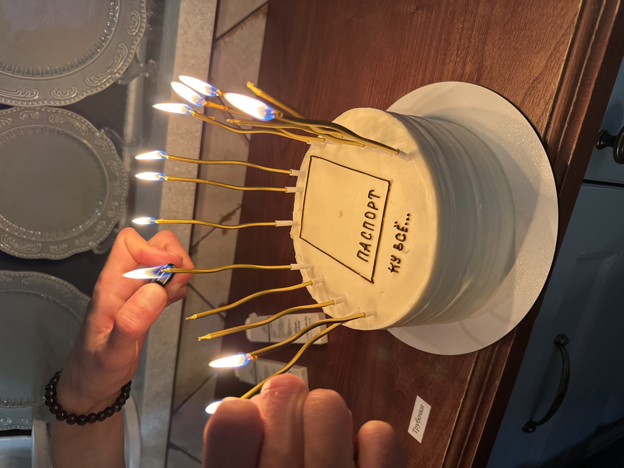 A person is lighting candles on a cake. The cake is white with a white plate underneath, and there is a rectangle drawn on the cake with the words "ПАСПОРТ" and "КУ ВСЁ..." written inside. The person lighting the candles has a bracelet on their wrist. A lighter can be seen in the person's hand. There are plates on the left and a wooden table on the right. A sticker with the word "Трубочки" is seen on the table. The location is likely not Sochi, Russia, as nothing in the image suggests that.