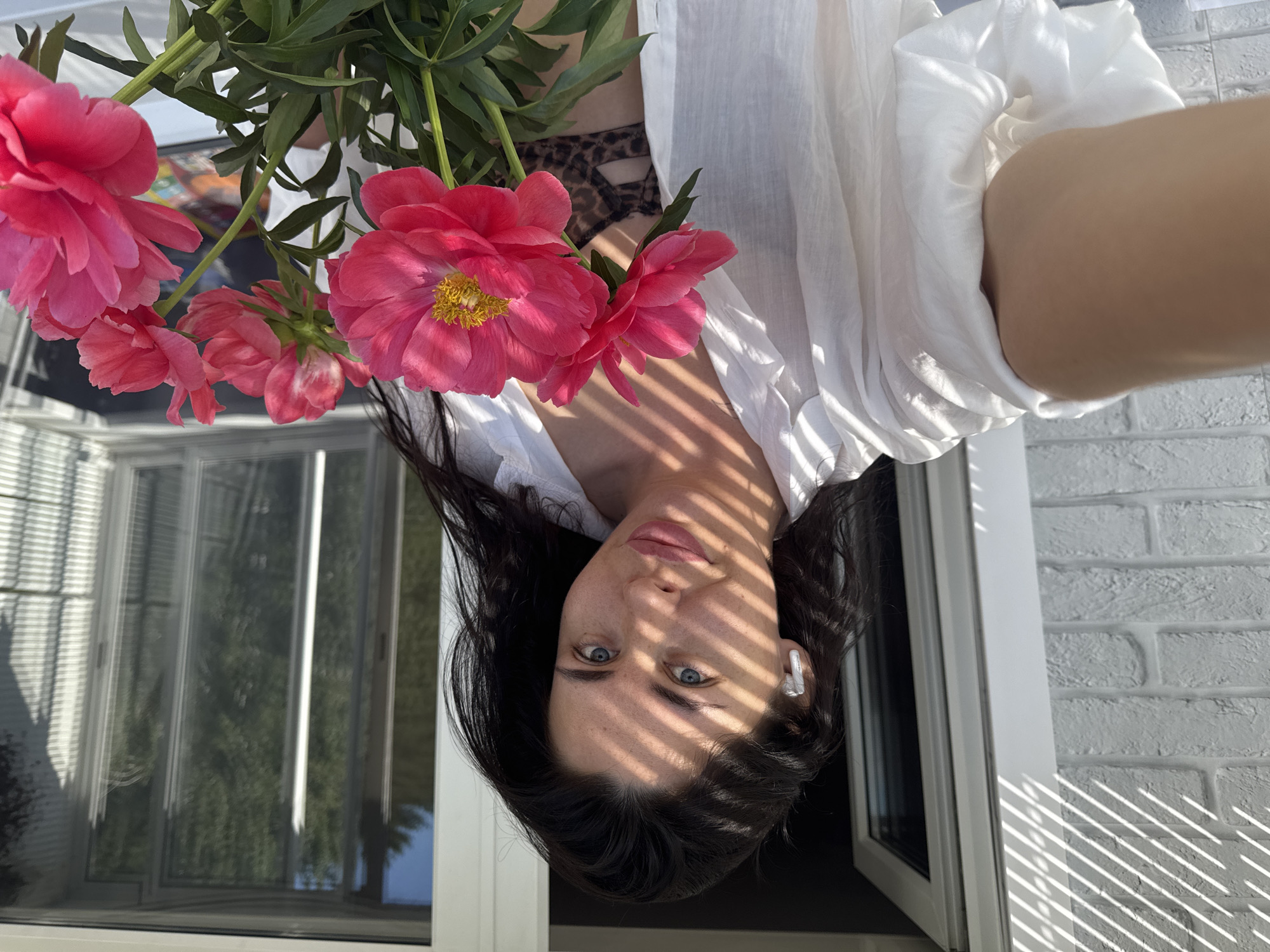 In Chaikovsky, Russia, a woman is pictured on a balcony with a white brick wall to her left and large glass doors and windows to her right. She is holding a bouquet of bright pink peonies and wearing a white unbuttoned shirt over a leopard-print bra. The sunlight casts striped shadows across her face and chest. She has dark hair and is wearing white earbuds. The scene appears to be outdoors during daylight.