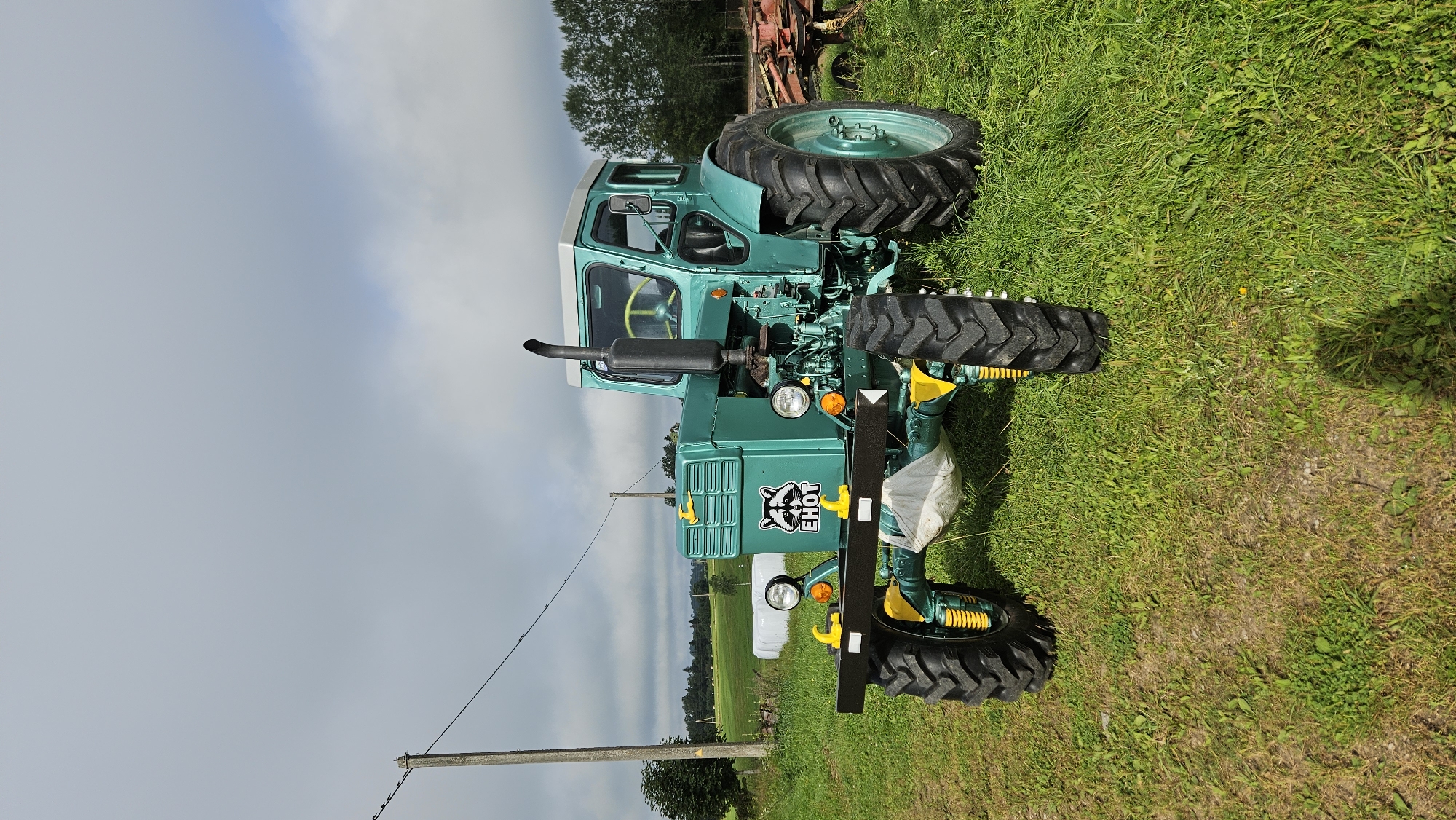 A turquoise vintage tractor with a black exhaust pipe and yellow accents is parked on a grassy field in Kapūņi, Latvia. The tractor has a stylized raccoon logo with the word "EHOT" below it on its front grille. In the background, there are trees, a utility pole, and a hazy sky. Some farm equipment can be seen to the right of the tractor.