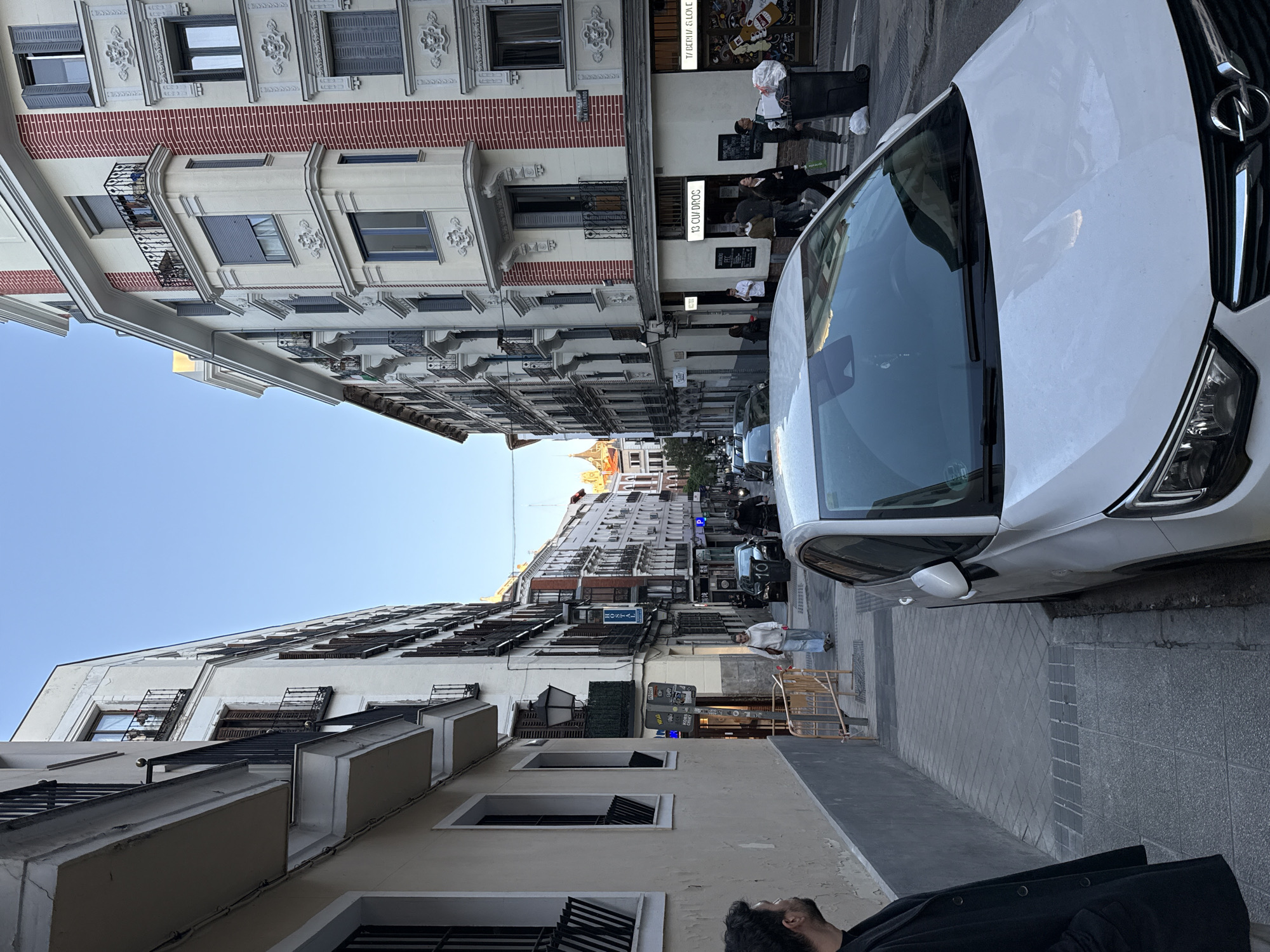 A ground-level, tilted photograph depicts a narrow street scene in Madrid, Spain. The street is lined with buildings of light-colored stone and brick, with multiple stories, balconies, and window shutters. The sky is visible between the buildings, showing a clear blue. A white Opel car is prominently parked on the right side of the street, partially obstructing the view down the street. Several people are visible along the sidewalk on the right, some standing in front of what appears to be a restaurant or bar. Other cars are parked further down the street. In the foreground, along the bottom edge of the frame, part of a person's head and shoulder are visible. The street is paved with gray stone.
