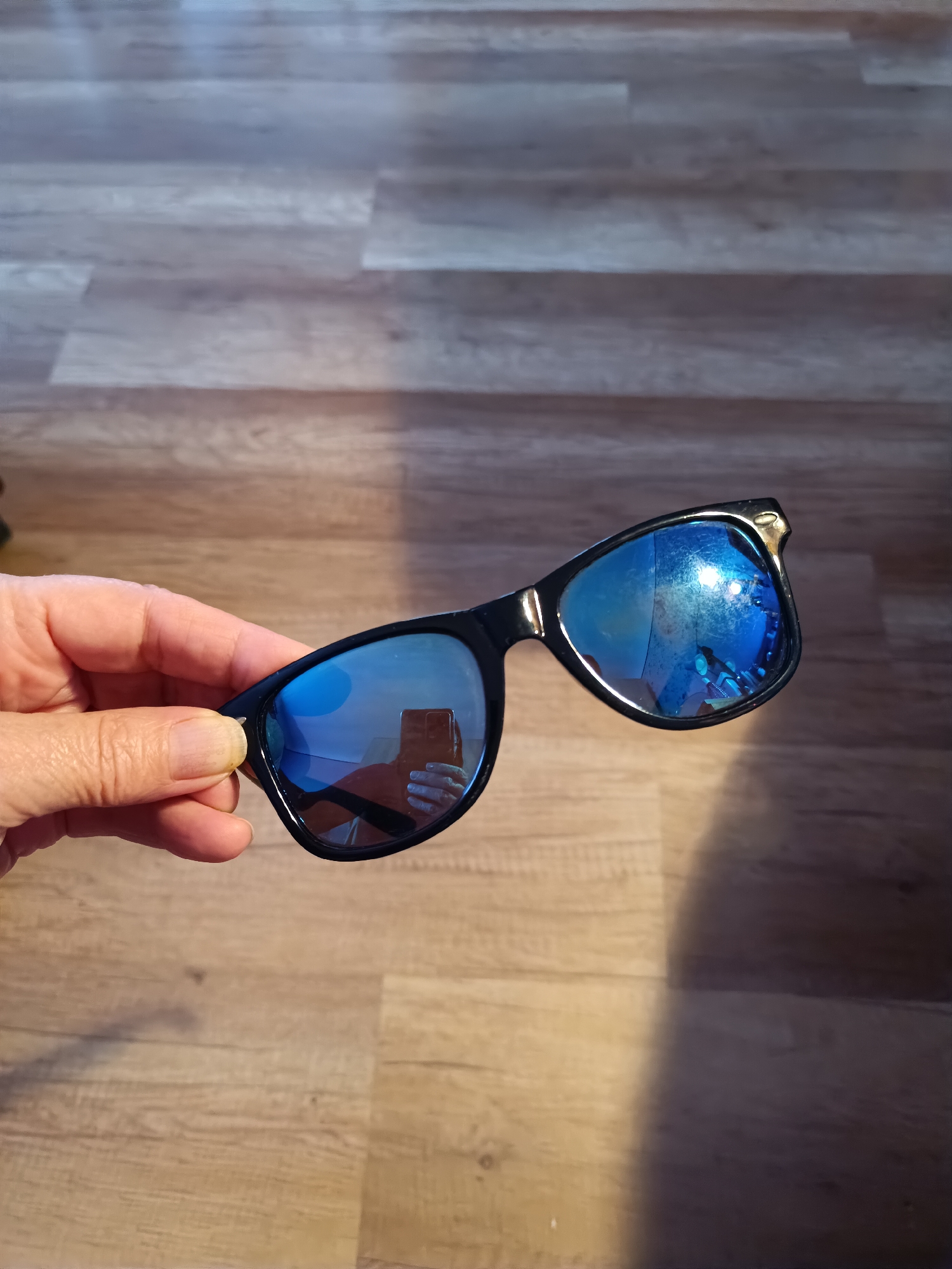 A person's hand holds a pair of black-rimmed sunglasses with blue mirrored lenses. The sunglasses are positioned in the foreground, with the hand holding them from the left side. The lenses reflect a bright blue sky, along with a hint of a person's face and a mobile phone being used to take the photograph. The background consists of a wooden floor with a light brown, planked pattern. The scene is illuminated, casting a shadow of the sunglasses and hand onto the floor. The image does not provide any information about the location in the city of Tebras, Latvia.