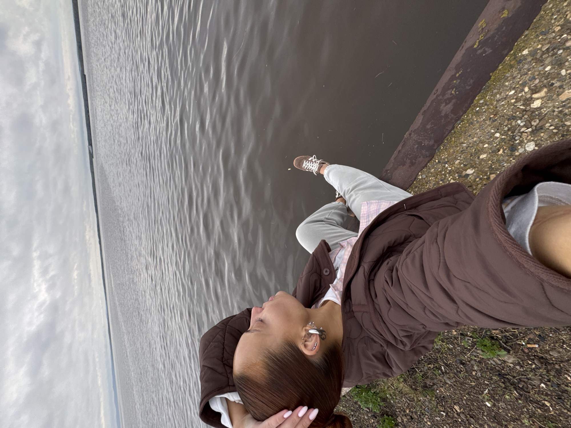 A young woman sits on a concrete embankment next to a body of water. The location appears to be Chastyye, Russia, based on the surrounding environment. She is wearing grey pants, a white shirt, a pink and white plaid shirt, and a brown quilted jacket. One leg is extended over the water, with a brown sneaker visible. She is looking towards the water and holding her hair. The water is a dark color, and the sky is overcast. The embankment is surrounded by small rocks and sparse vegetation.