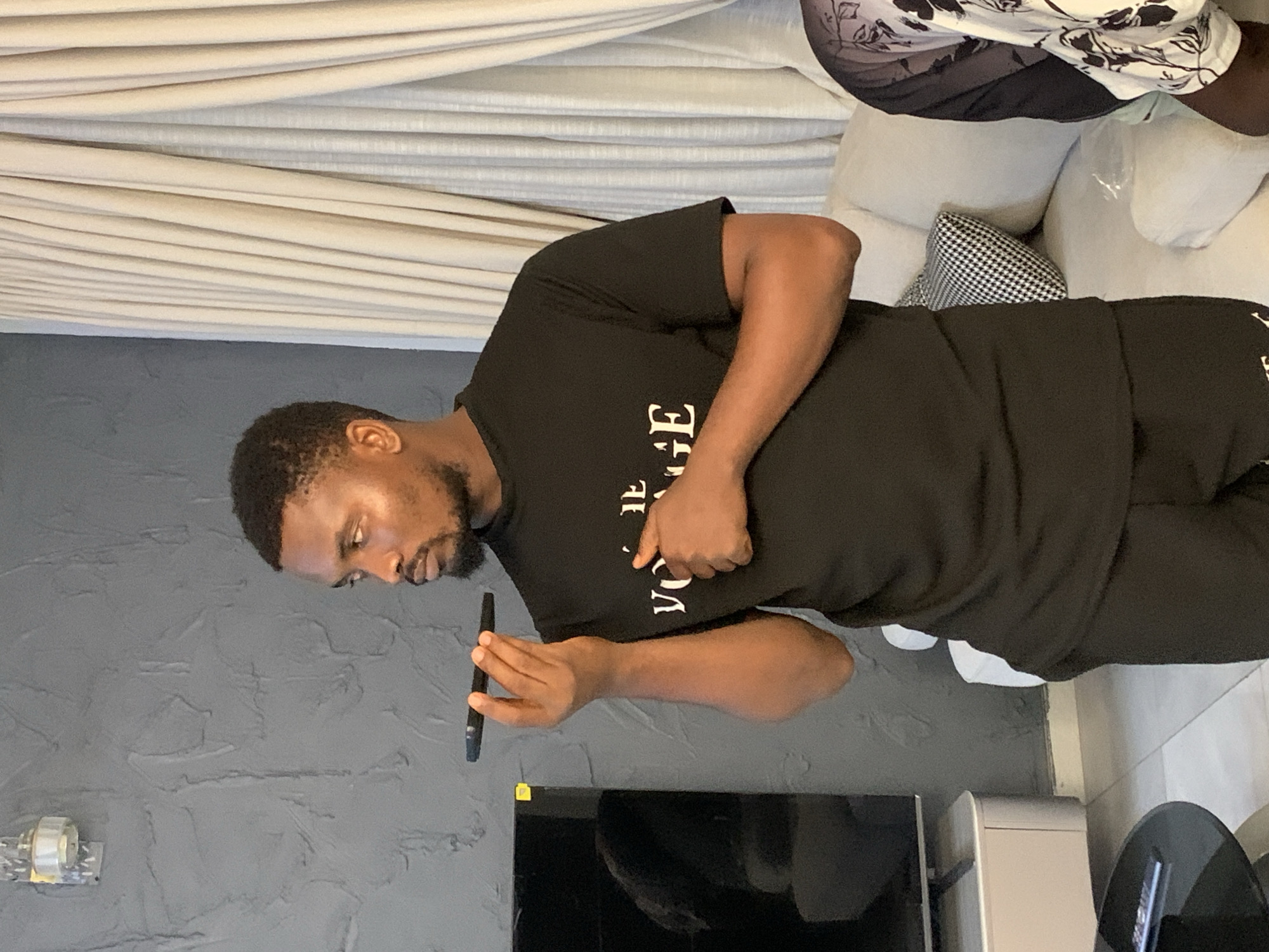 The image depicts an indoor scene in Igboefon, Nigeria. A dark-skinned adult male with short dark hair and a beard stands centrally, wearing a black short-sleeved t-shirt with partial white text ("THE VOYAGE") and black trousers. He holds a black smartphone horizontally to his mouth with his right hand, suggesting interaction with the device's audio functions, while his left hand rests on his chest. His gaze is directed towards the right. To his right, a second individual is partially visible, seated on a light-colored sofa and wearing a patterned garment. The background features a textured dark grey wall with a small wall-mounted light fixture in the upper left. Light-colored, pleated curtains are visible on the right side of the frame. A large, inactive flat-screen television rests on a light-colored media console in the left background. A black, circular coffee table is partially visible in the lower left foreground, and the floor consists of light-colored tiles.