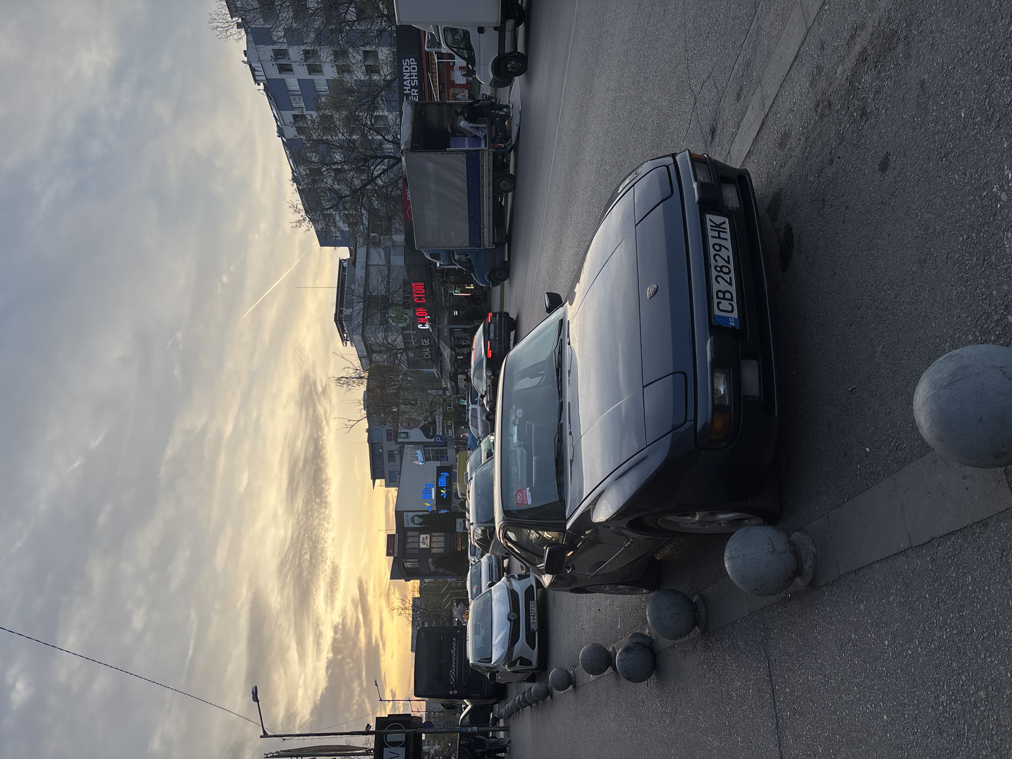 This outdoor image captures an urban street scene in Sofia, Bulgaria, during late afternoon or sunset, indicated by the warm, golden light filtering through the clouds in the sky. The perspective is slightly elevated, looking down a street lined with buildings and vehicles.
In the foreground, a dark grey/blue classic sports car, possibly a Porsche, is parked on the asphalt. Its Bulgarian license plate, "CB 2829 HK," is clearly visible, along with the "BG" country sticker. A row of grey, spherical concrete bollards runs along the curb next to the parked cars.
Further down the street, a line of other vehicles is parked, including a white Renault with the Bulgarian license plate "CB 6497 CO." A large white truck is visible further down, with people standing near its open rear. A black bus or truck featuring the text "BARBAROS" on its side can also be seen.
The street is flanked by multi-story buildings, appearing to be a mix of residential and commercial structures. Some buildings have balconies, and bare tree branches are visible, suggesting a colder season. The cloudy sky in the upper left shows a distinct contrail.
Visible text on various signs includes "HANDS ER SHOP" (likely part of "BARBER SHOP"), "CASH CTOP" (likely "CASH STOP" or similar money exchange in Cyrillic), "dm" (a drugstore logo), and "Filly." The scene depicts a typical bustling city street during the evening hours.
