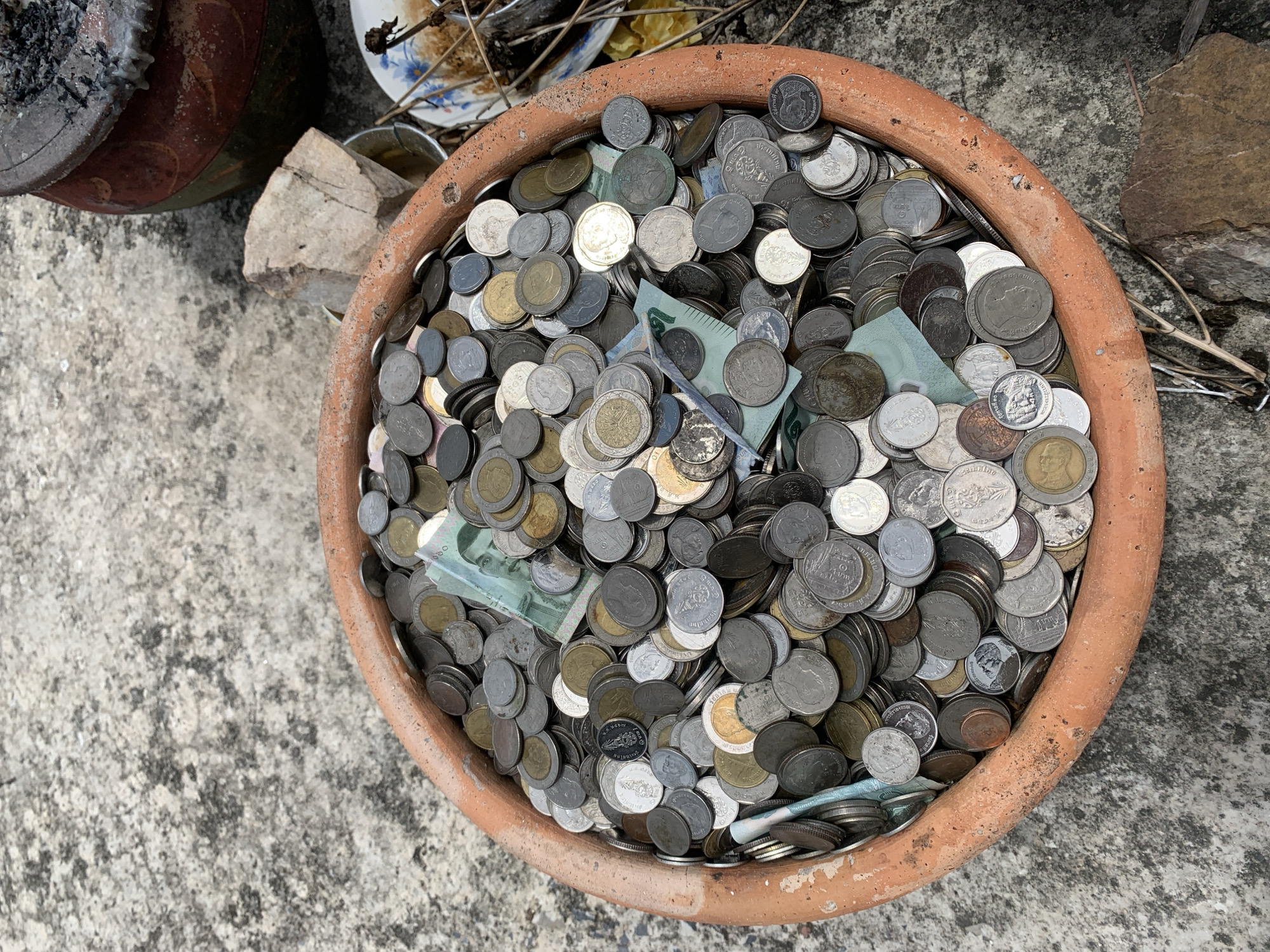 A clay pot overflowing with a large quantity of coins and some Thai baht banknotes is positioned on a concrete surface in an outdoor environment. The pot is terracotta-colored and appears to be roughly circular. The coins are of various sizes and metallic colors, including silver, gold, and some with a darker hue. Several banknotes, appearing to be light green in color with images of a person, are visible amongst the coins. To the right of the pot, a small portion of a rustic, dark brown ceramic vessel with ashes or embers is visible, suggesting it may be a brazier or incense burner. A light-colored, rough-textured rock sits next to the pot. In the background and to the right, there is a partially visible white plate with a floral pattern, containing some dried plant matter and what looks like food remnants. The concrete ground is textured with patches of discoloration and wear. The scene appears to be located in an outdoor setting in an unspecified area of Thailand.