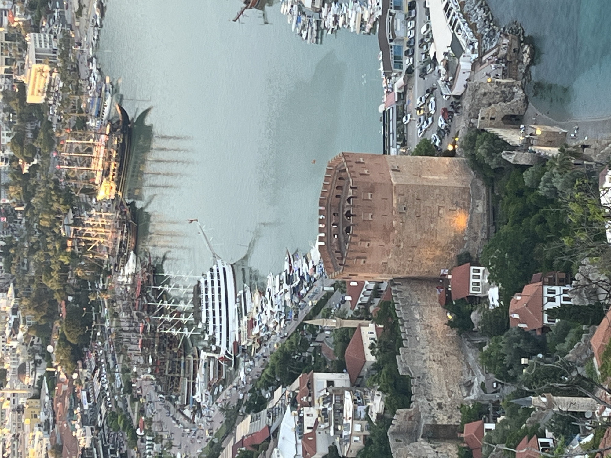 The image captures an elevated view of Alanya, Turkey, likely from a high vantage point on the city's peninsula. Prominently featured is the Kızılkule (Red Tower), a historical tower made of red brick, located near the harbor. The harbor area includes a mix of modern yachts and traditional wooden ships. The urban landscape around the harbor is densely built with residential buildings. The sea occupies a significant portion of the image, and the light suggests it is either sunrise or sunset. The ancient city walls meander through the landscape.