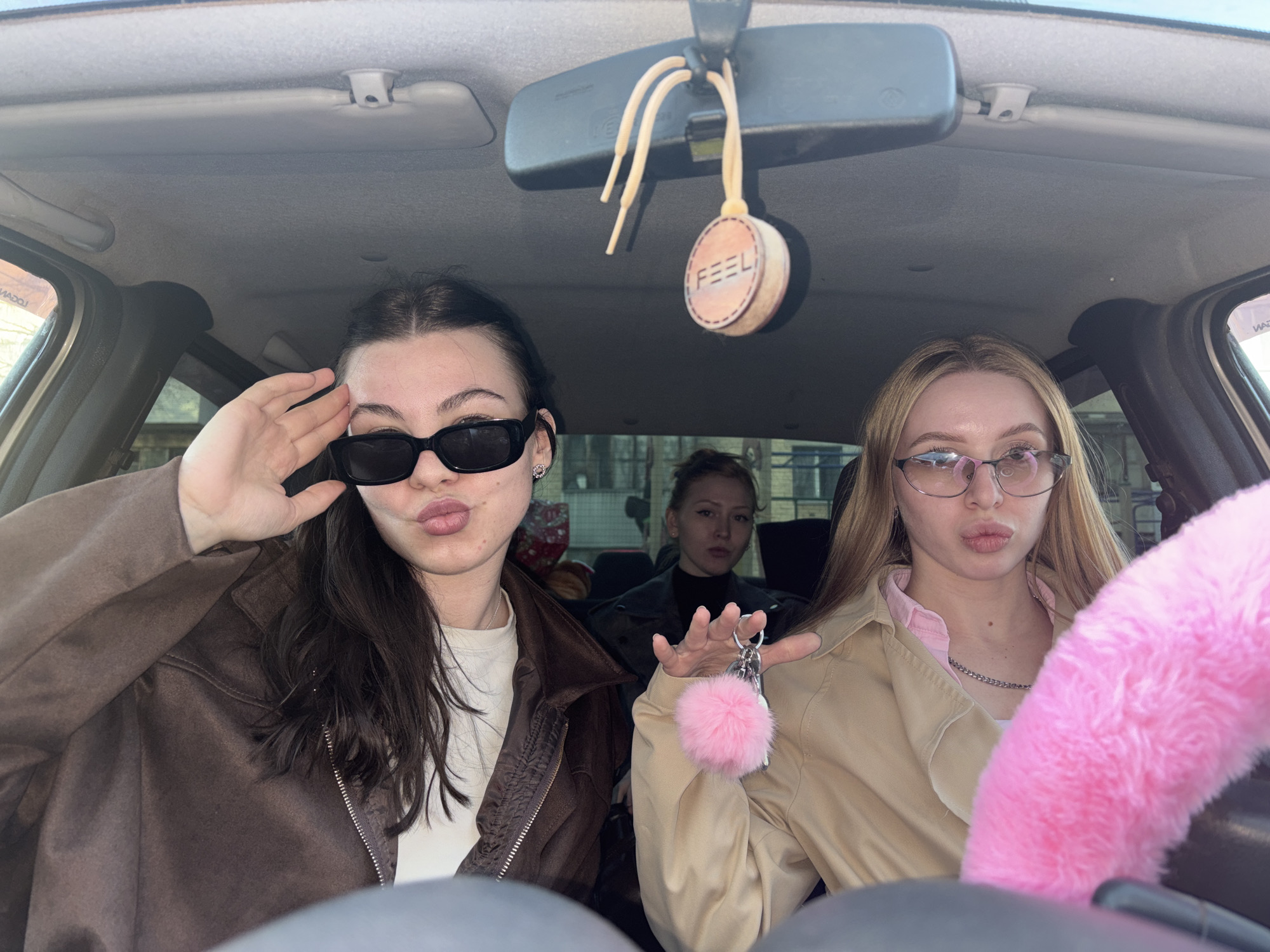 Three young women are captured in a selfie-style photograph taken from within a car's interior during what appears to be a bright day. The setting is inside a passenger vehicle, suggested by the rearview mirror, sun visors, and parts of the dashboard and steering wheel. The specific location is Perm, Russia.
In the front left, a woman with dark hair wears sunglasses, a brown jacket over a white top, and a subtle earring. She makes a kissy face and raises her right hand to her forehead in a salute-like gesture. To her right, in the front passenger or driver's seat, a woman with blonde hair and glasses wears a beige jacket over a pink collared shirt and a silver chain necklace. She also makes a kissy face and holds a set of car keys adorned with a pink fluffy pom-pom in her left hand. A prominent pink furry steering wheel cover is partially visible in front of her. In the back seat, a third woman with dark hair pulled back looks towards the camera with a neutral expression.
Visible car accessories include a circular, tan-colored air freshener or ornament hanging from the rearview mirror, which has the word "FEEL" embossed on it. The text "LOGAN" is visible in reverse on a sticker or label on the side window, suggesting the car might be a Dacia/Renault Logan model. The overall mood is playful and casual, typical of friends posing for a photo. The clear lighting indicates it is daytime, and the visibility through the windows suggests fair weather.