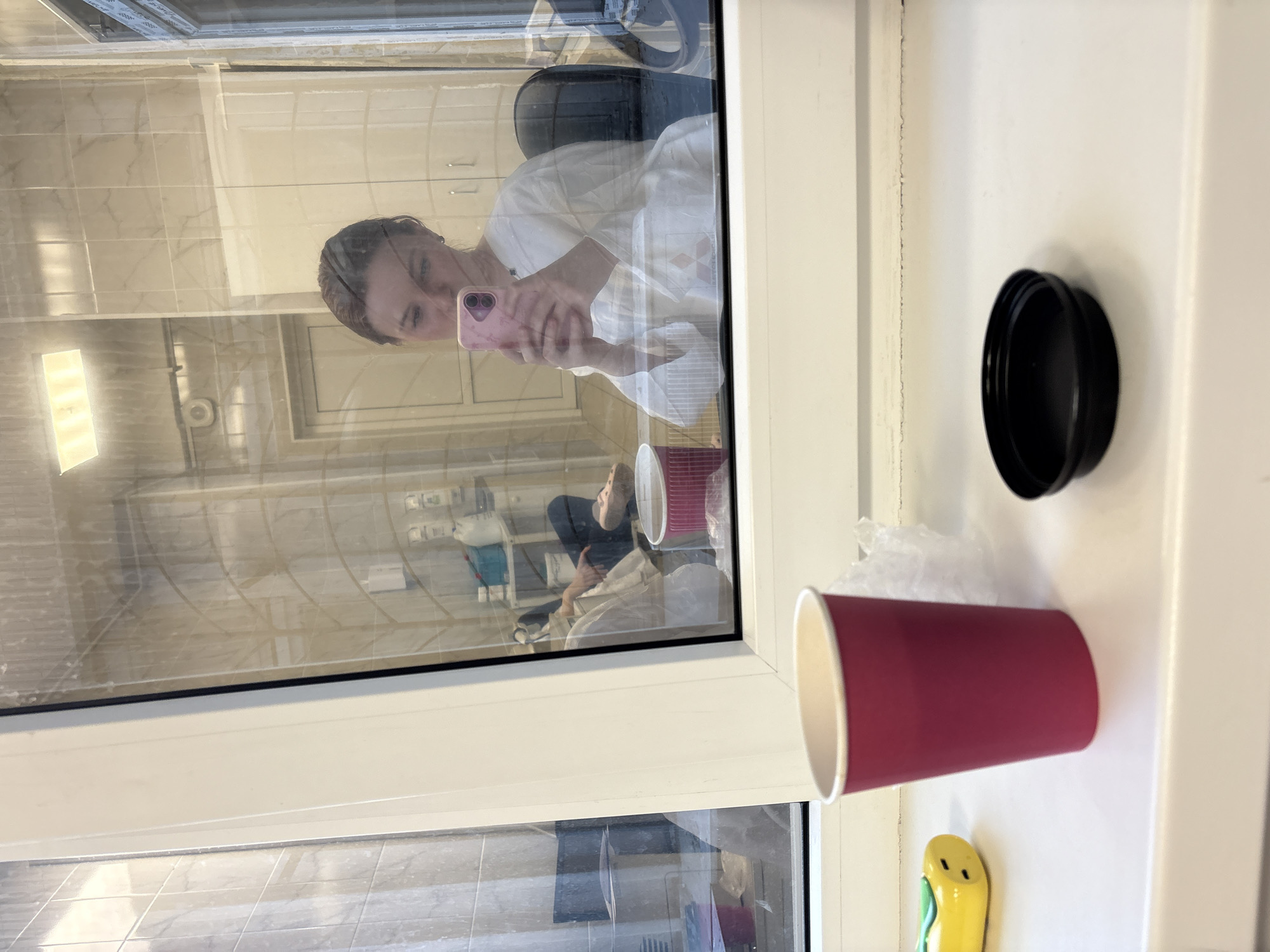 The image shows an interior scene viewed through a window in what appears to be Saint Petersburg, Russia, based on the presence of specific window styles commonly found in the city. The reflection in the window shows a person holding a pink mobile phone, presumably taking the picture. The person is sitting in what appears to be an office or living space, with a desk chair visible in the background. Behind the person, the reflection shows what appears to be another individual sitting on a chair, possibly in a bathroom setting given the tiled walls and fixtures in the reflection. A red disposable cup and a black lid sit on the windowsill in the foreground, along with what appears to be crumpled plastic wrap and a small yellow object, possibly a cleaning tool. The window frame is white. The tiles visible through the reflection look typical for apartment buildings in this city.