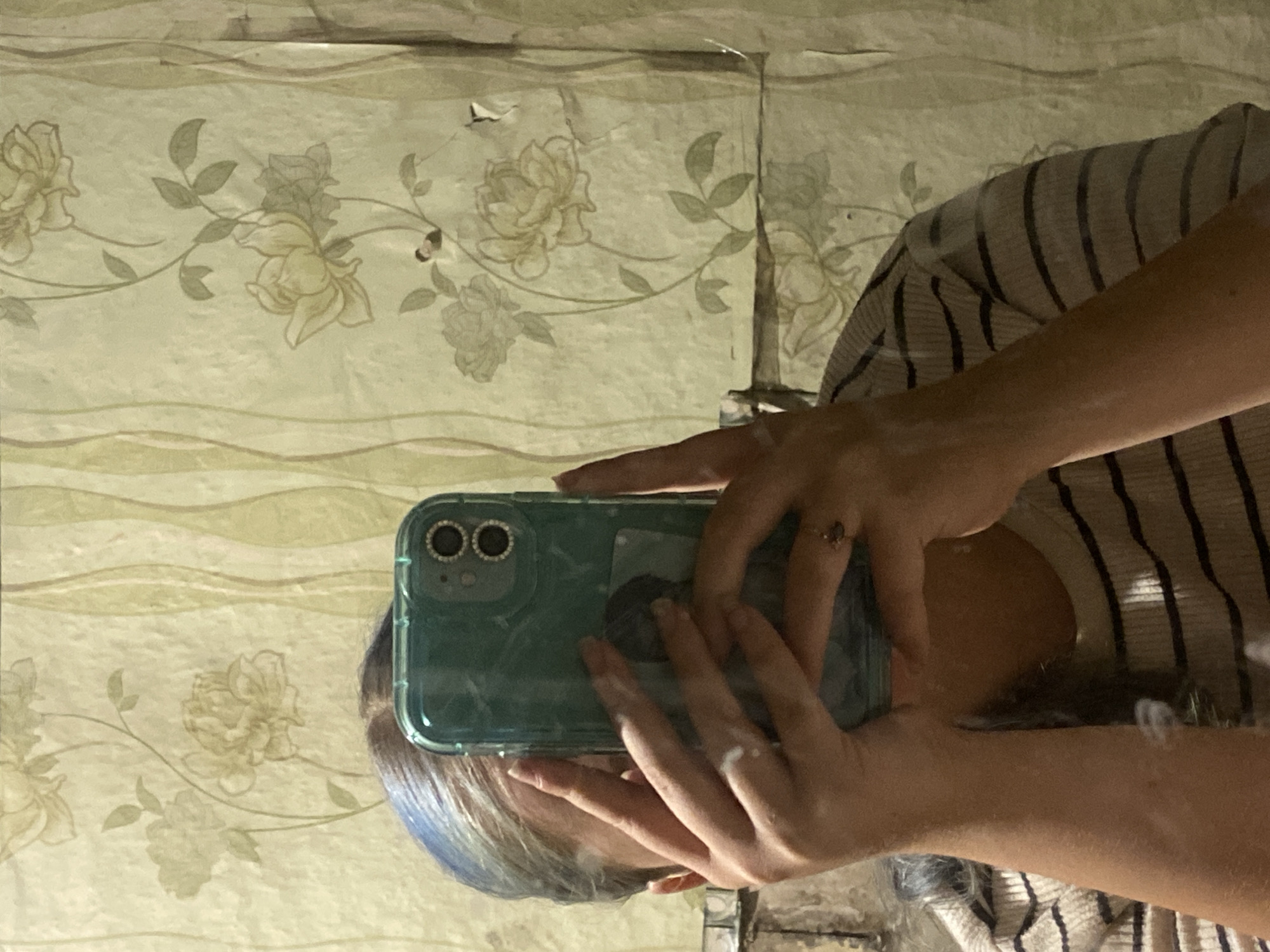 A young woman with blue-tinted hair takes a selfie in a bathroom mirror. She is wearing a striped t-shirt and holding a teal-colored phone with a marble-patterned case. The background is a faded floral wallpaper with a slightly torn texture. The lighting is dim and casts a shadow on the right side of the image. The image was taken in Nova Praha, Ukraine.