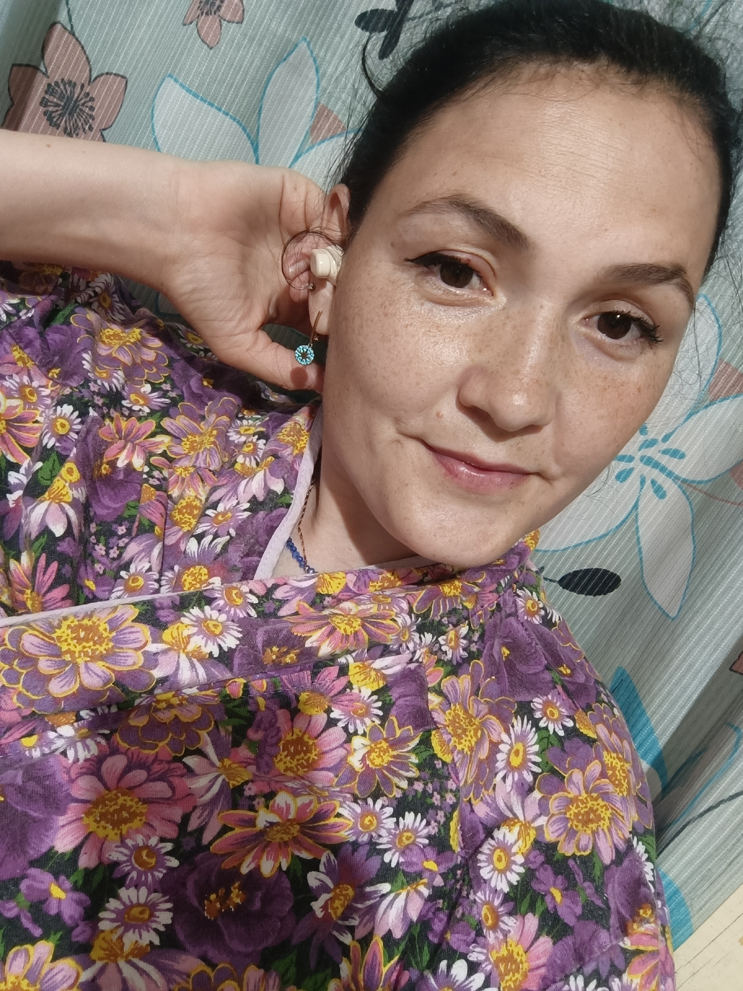 The image is a close-up selfie of a woman with dark hair and fair skin, adorned with freckles. She is wearing a purple floral-print top and has her left hand raised, touching her ear where a small earring with a blue gem is visible. She is also wearing an earbud in her left ear. The background consists of a curtain with a floral pattern in shades of blue, green, and white. The setting is indoors, likely a home environment. There is no indication of the city of Karsha, Russia in the image.