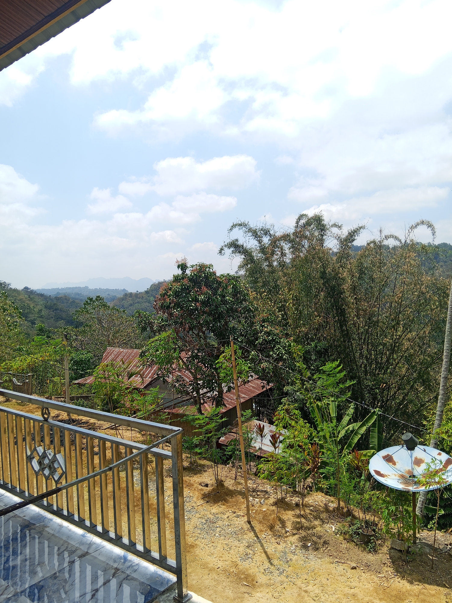 The image depicts a serene landscape in Batu Papan, Indonesia, featuring a view from a balcony. In the foreground, a metallic railing with a decorative diamond pattern runs along the edge of the balcony, which has a tiled floor. Beyond the railing, a sloped terrain is visible, covered with dry leaves and scattered stones.
Mid-ground, several structures are nestled among lush greenery. Two houses with corrugated metal roofs are partially visible, suggesting a rural or semi-rural setting. A satellite dish, rusted and weathered, stands prominently on a metal pole to the right, surrounded by dense vegetation. Various types of trees and plants, including bamboo and banana trees, contribute to the verdant environment.
The background reveals a rolling, mountainous terrain covered in dense forest. The distant mountains fade into a hazy horizon under a sky with scattered white clouds. The overall impression is one of a peaceful, natural setting with signs of human habitation.