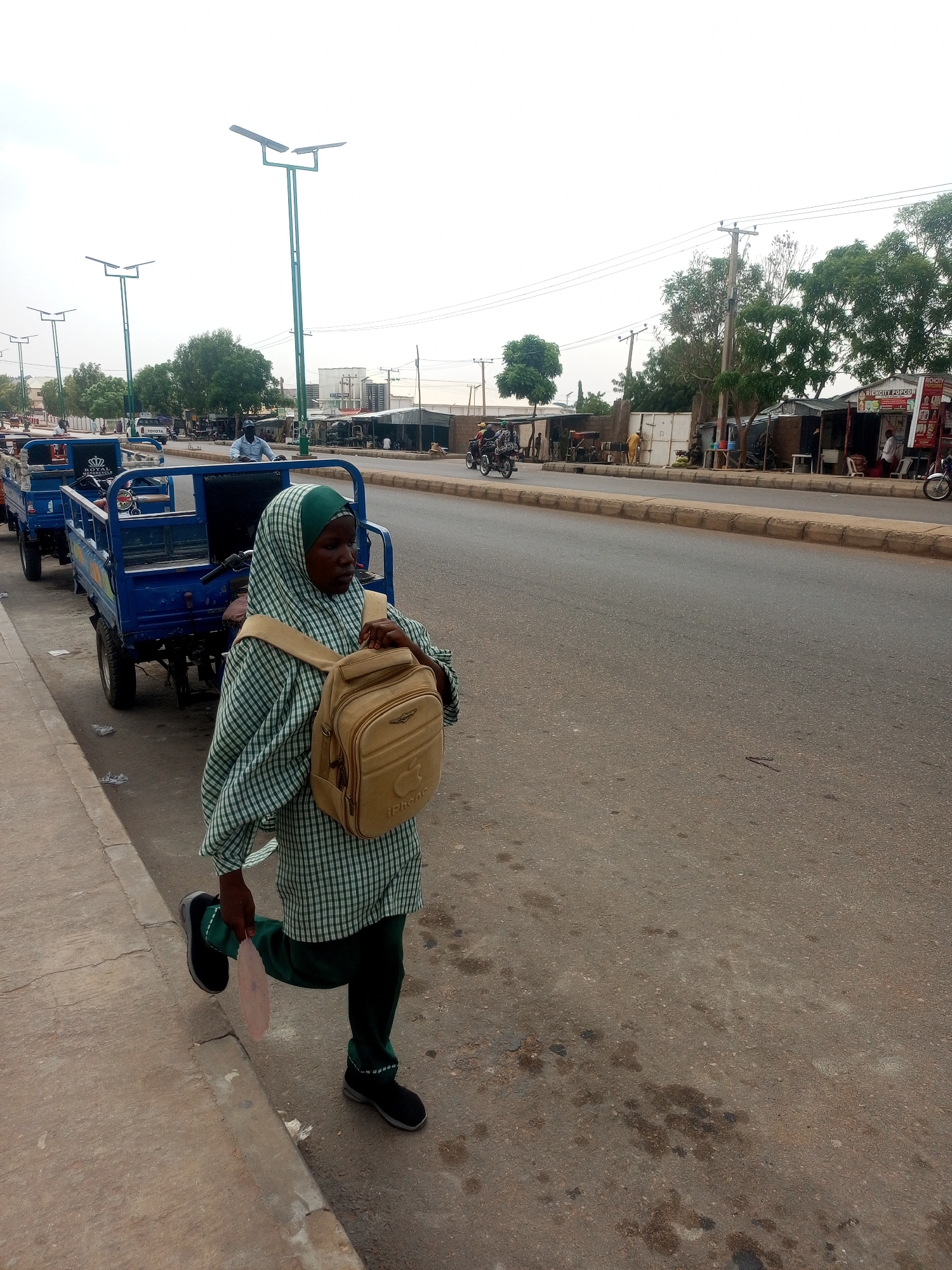A person wearing a green and white checkered top, dark green pants, black shoes, and a tan backpack walks along the side of a street in Unknown, Unknown. The person is holding a round, light-colored object in their left hand. The person is wearing a green head covering. The street is wide and made of asphalt. Blue three-wheeled vehicles are parked along the curb to the left. Streetlights are visible in the background. Buildings and trees line the far side of the street. Motorcycles are visible on the street. Overhead utility lines are present. The sky is overcast.