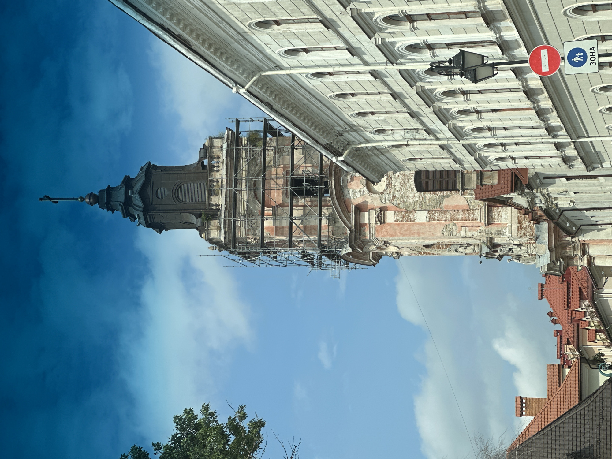 The image captures a view of the Dominican Church and Monastery in Kamianets-Podilskyi, Ukraine. A tall, weathered bell tower, partially covered in scaffolding, dominates the left side of the frame. The tower displays signs of age and renovation, with exposed brickwork and crumbling plaster. Adjacent to the tower, on the right, is a light-colored building with a facade featuring numerous arched windows. A black lamppost with two lamps stands in front of this building, accompanied by two traffic signs: a red circular sign with a white horizontal bar and a blue square sign depicting two figures, one adult and one child, above the text "ЗОНА". The sky above is a vibrant blue with scattered white clouds. In the foreground, to the left, are the red-tiled roofs of other buildings. The scene appears to be an urban street view under daylight conditions.
