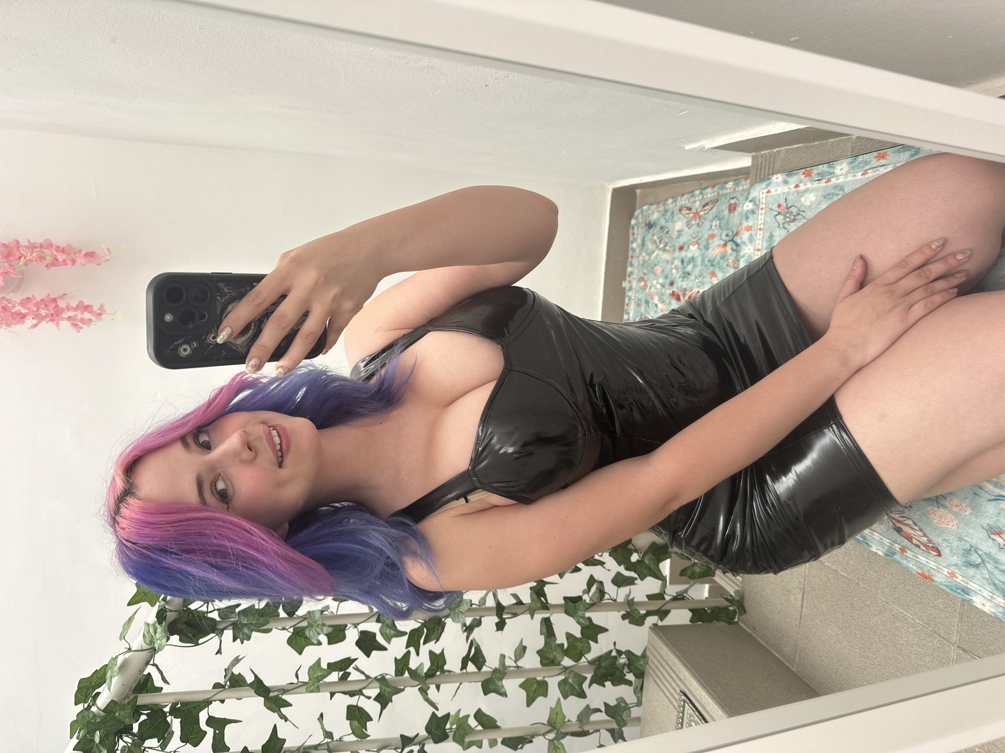 A young woman is captured in a mirror selfie, holding a black smartphone. She has long, vibrant hair, styled with pink strands framing her face and blending into blue/purple hues towards the ends. She is smiling faintly, looking directly into the camera. Her nails are manicured with a glittery design.
She is wearing a form-fitting, short, black dress made of a shiny material, possibly latex or PVC, with a bustier-style top. Her left hand holds the phone, while her right hand rests casually on her thigh.
The setting appears to be an indoor space, possibly a room or hallway. To her left, behind her, there are artificial pink flowers attached to a white wall. Below them and extending across the lower left of the frame is a white railing or lattice adorned with green artificial ivy. The background also reveals a light blue/teal rug with a pattern of butterflies and floral motifs, and a tiled surface below it, likely a floor or lower wall. The lighting suggests it is daytime.
No text is visible in the image. The provided location context is the City of Zagreb, Croatia, but this is not identifiable from the image itself.