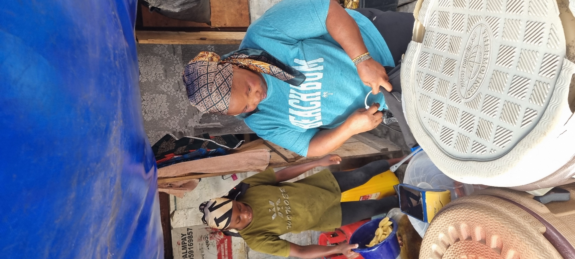 Two women are standing under a blue tarp, possibly at a market stall in Lagos, Nigeria. The woman on the left, wearing a green t-shirt and headscarf, is holding a blue bucket filled with yellow food items, possibly plantains, and appears to be pouring them into a larger container. The woman on the right, wearing a turquoise t-shirt and a patterned headscarf, is looking down and using her hands to open a black bag with a white zipper pull. In the foreground, the soles of two large shoes or footwear are visible. Behind the women, there are various items including what looks like a red gas cylinder, fabric, and a notice on a wall that reads "PALMPAY 059169857." The environment suggests an outdoor setting, likely a marketplace, with makeshift shelter.