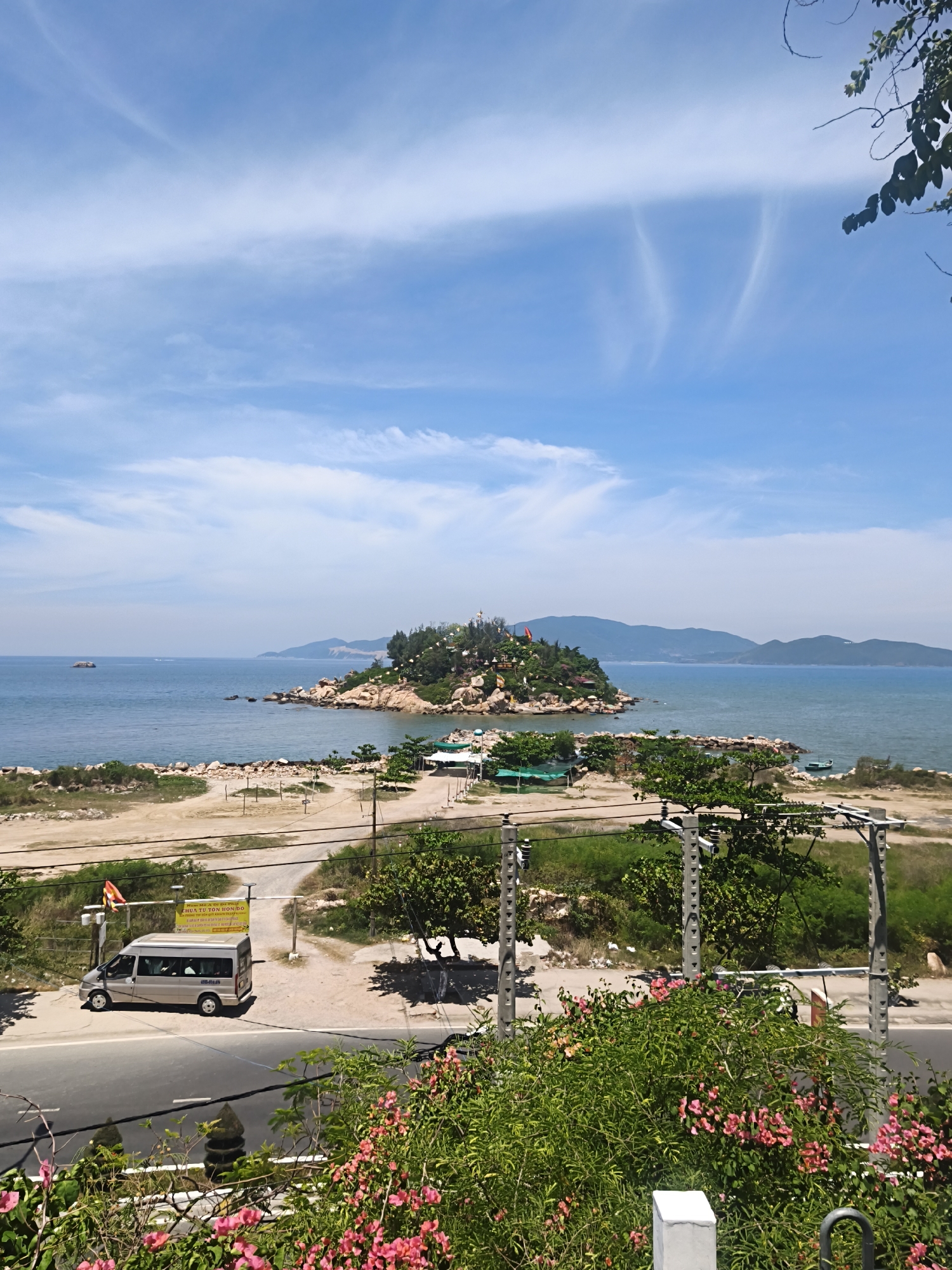 This is a scenic view of the coastline in Nha Trang, Vietnam. A small, rocky island dotted with trees and structures is situated in the ocean. In the foreground, a road runs parallel to the coast, with a silver van parked on its shoulder. Lush green vegetation and pink flowers bloom on the right side of the image, framing the scene. The sky above is a clear blue with scattered white clouds, and distant hills can be seen on the horizon. The overall impression is one of a peaceful coastal landscape.