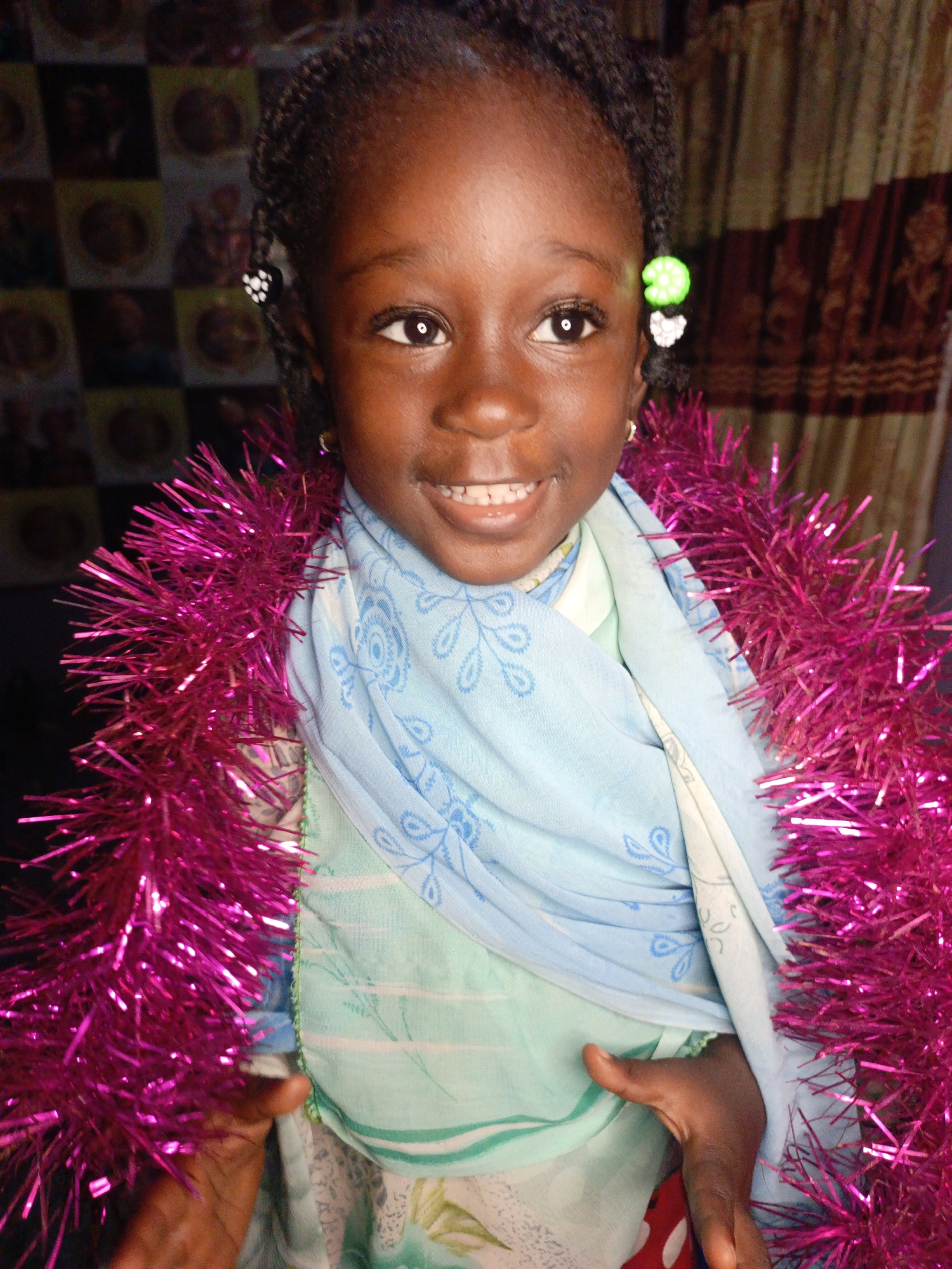 The image shows a close-up of a young Black girl in Akwanga, Nigeria, wearing a bright pink tinsel garland around her neck and a light blue patterned scarf over a light green garment. She has braided hair, decorated with black and white beads and a green charm. The background consists of a patterned curtain and a wall with multiple framed images. The girl is smiling, with her teeth visible. Her right hand is visible, partially covered by the tinsel. The lighting suggests an indoor setting.
