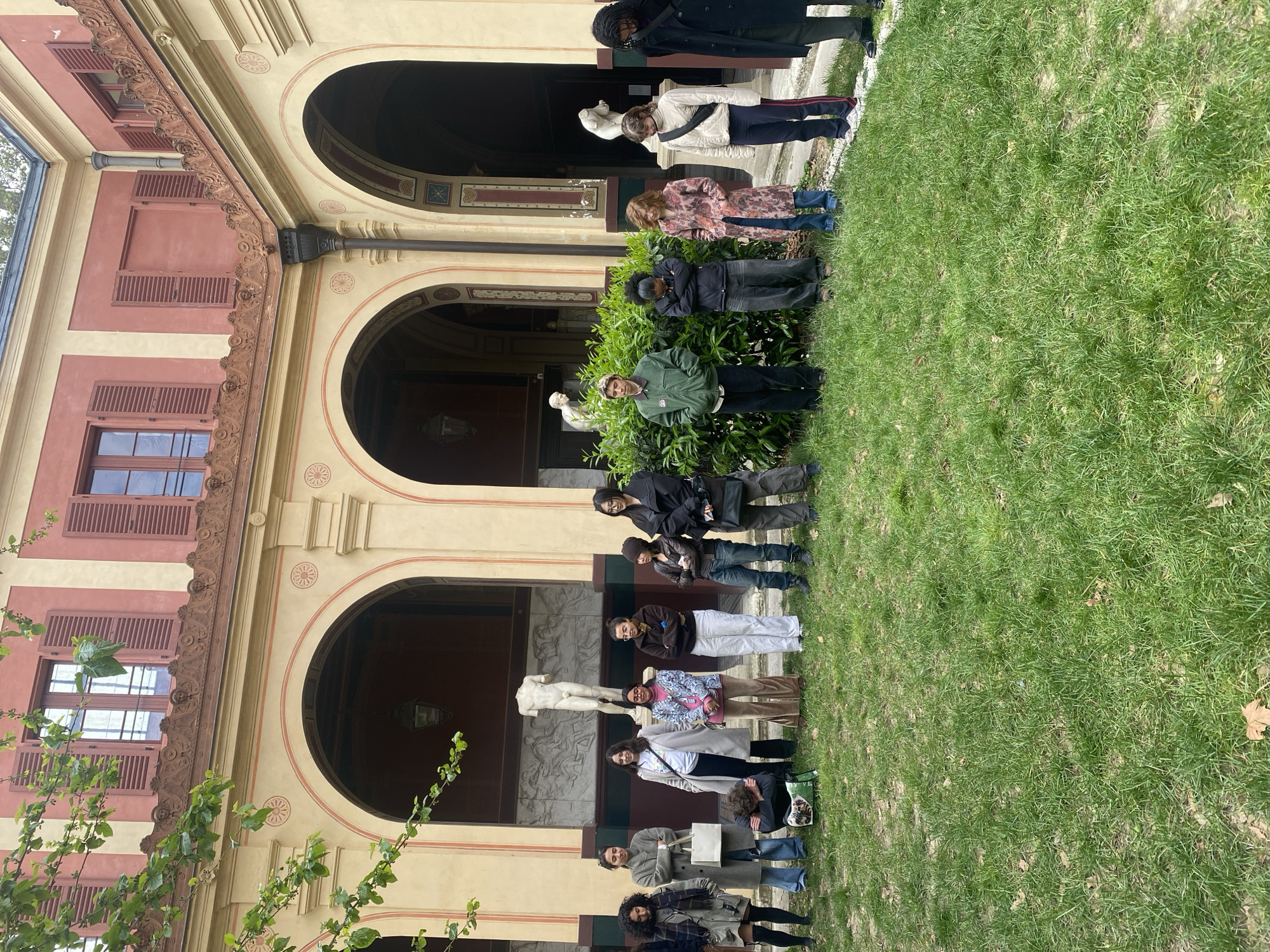 A group of approximately 15 people stands on a grassy lawn in front of a building with arched openings. The building has a façade of pink and tan hues with decorative architectural details and windows with brown shutters. Statues can be seen through the arched openings. The image appears to have been taken at the Musée Rodin in Paris, France, given the architectural style and statues. The people are varied in age and dress and appear to be posing for a group photo.