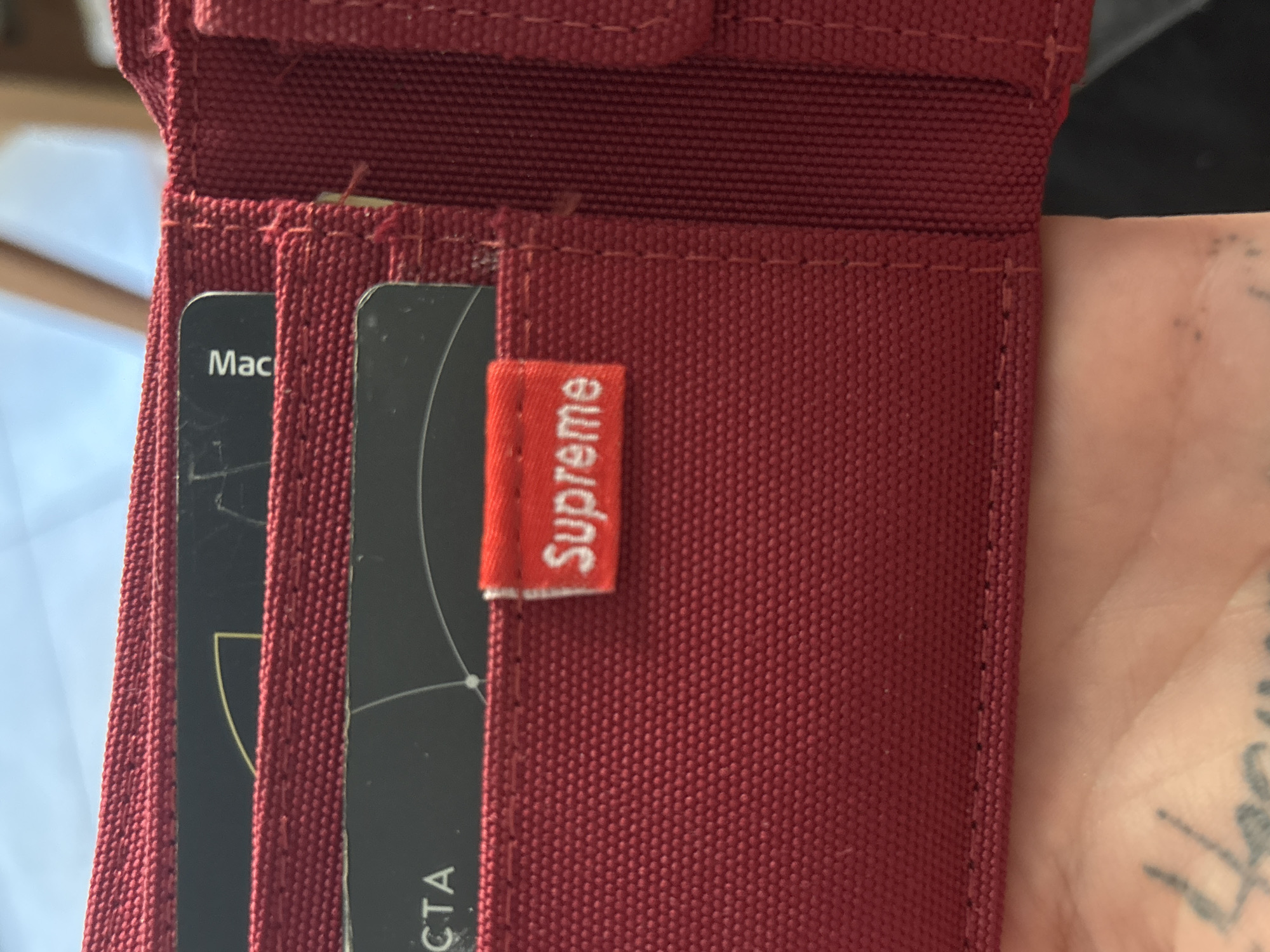 A close-up shot reveals a maroon wallet with a red "Supreme" tag on one of the card slots. Two cards are partially visible in adjacent slots. The card on the left is black with "Mac" visible, and the card on the right is black with "CTA" visible. A tattooed hand is visible in the background. The setting is unknown.