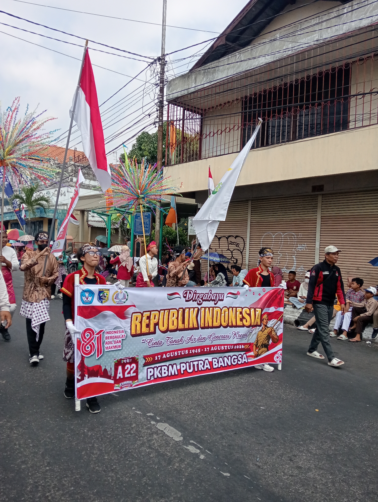 In the city of Bintoro, Indonesia, a parade is taking place on a paved street under an overcast sky. The parade features a group of people, some of whom are dressed in traditional Indonesian attire, carrying flags and a large banner.
The banner prominently displays "Dirgahayu Republik Indonesia" in large, bold lettering, indicating a celebration of Indonesian independence. Below this, smaller text reads "Cinta Tanah Air dan Generasi Kreatif," translating to "Love the Homeland and Creative Generation." The banner also includes the dates "17 Agustus 1945 - 17 Agustus 2026," marking the 81st anniversary of Indonesian independence. A depiction of a man holding a flag is featured on the right side of the banner, accompanied by the text "PKBM PUTRA BANGSA."
Several individuals are actively participating in the parade. In the foreground, a young person wearing glasses and a red and black traditional top is holding the banner with white gloves. To their left, another participant in a gold traditional outfit and a patterned skirt holds a long pole. Further back, other participants are visible, some with colorful, fan-like decorations mounted on poles. Two Indonesian flags, red and white, are also being carried.
Spectators line the street, seated and standing on the sidewalk, watching the procession. The background shows buildings with windows and some shops with closed shutters. Utility poles and wires are visible overhead, typical of an urban street scene. The overall atmosphere suggests a festive and patriotic event.