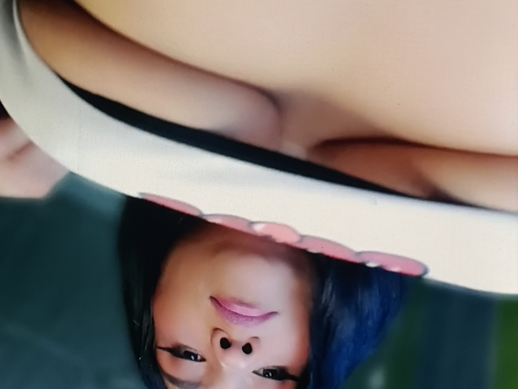A person's face is angled downwards and to the left, with dark hair framing their face. They have dark eyes, a small nose, and full pink lips. They are wearing a white garment with a black trim and pink accents. The background is a blurry dark green.
