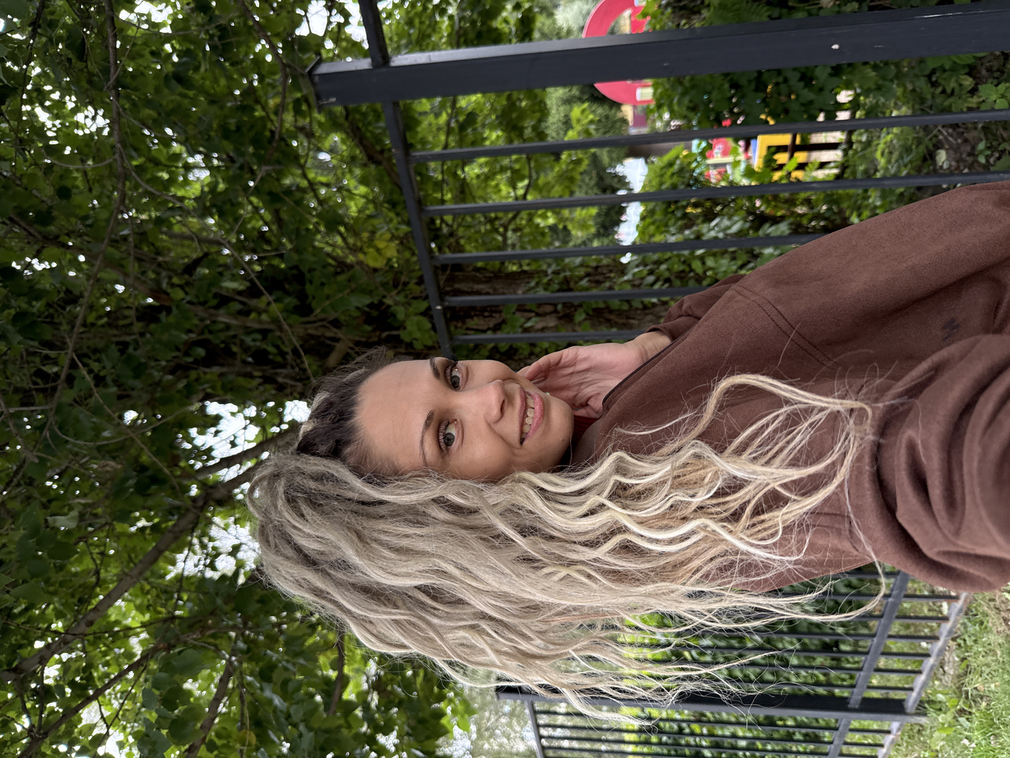 A medium shot captures a woman from the chest up, posed outdoors. She is facing left, with her head turned towards the camera and her right hand touching her face near her cheek. The woman has long, blonde, curly hair pulled back into a high ponytail. She is wearing a brown hooded sweatshirt. Behind her, a black metal fence runs horizontally across the frame, with a tree and foliage visible beyond it. Green grass covers the ground in the foreground. The background suggests an urban or park setting, with glimpses of colorful structures, possibly playground equipment, and more greenery. The lighting is natural, indicating daylight. The location is identified as Rusanovo, Russia.