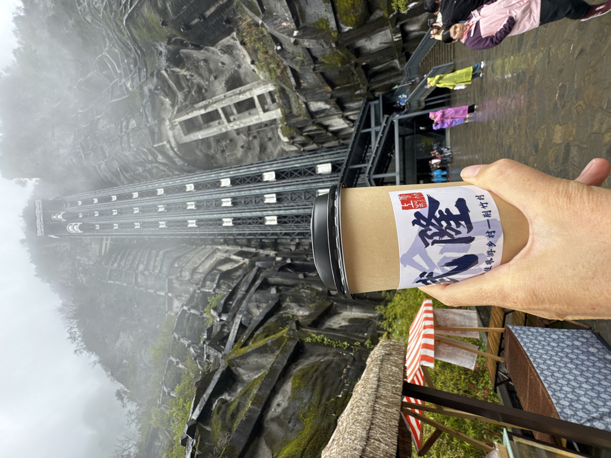 A hand holds a paper cup with a label in front of the Bailong Elevator in Wulong District, China. The elevator is constructed on a cliff face. Other people are visible. A small stall or kiosk is situated nearby. The sky is overcast.