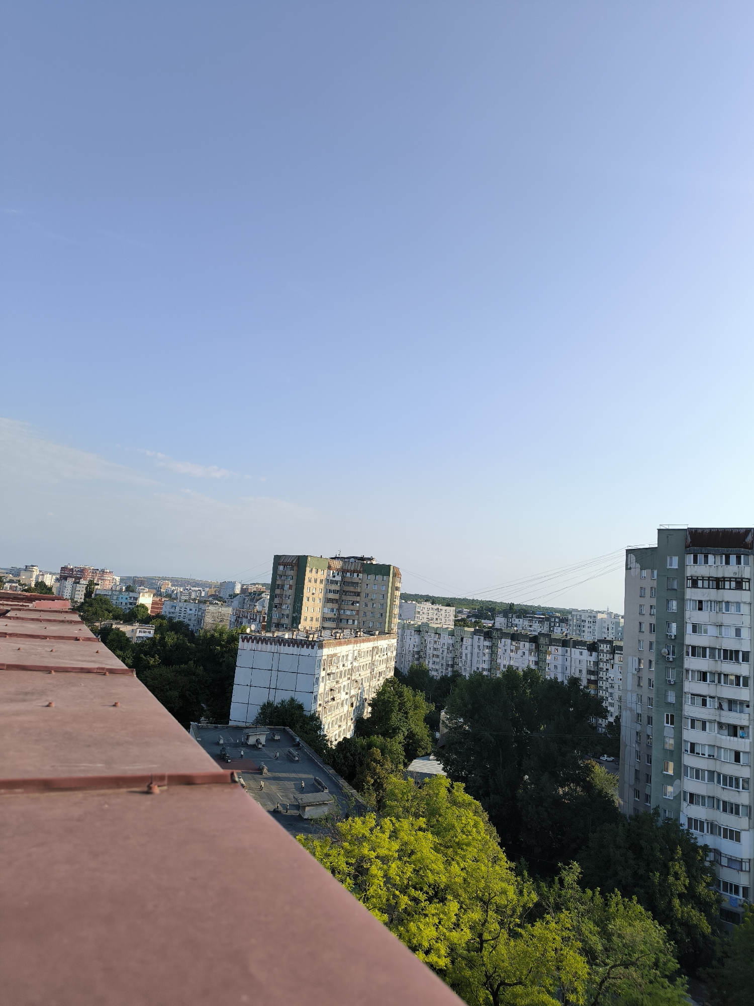 This is an outdoor, eye-level, full shot, landscape photograph taken from a high vantage point, possibly a rooftop, overlooking a city scene. The foreground is dominated by a large, reddish-brown metal roof with visible fasteners. Beyond this roof, the scene unfolds to reveal a dense urban environment characterized by numerous apartment buildings and trees.
The apartment buildings are of varying heights and architectural styles, suggesting a mix of older and perhaps more modern construction. Many of them appear to be of Soviet-era design, common in many Eastern European cities, with repetitive window patterns and light-colored facades. The buildings are clustered together, with some showing signs of wear and tear.
Interspersed among the buildings are numerous trees, some with lush green foliage, indicating a temperate climate and possibly a springtime or summer setting. The trees provide a significant amount of greenery, softening the harsh lines of the urban landscape. In the mid-ground, there's a visible line of power or telephone wires stretched between poles.
The sky above is a clear, pale blue with a few wispy clouds towards the left horizon, suggesting it is daytime and likely late afternoon or early evening due to the soft lighting. The overall impression is of a densely populated urban area with significant tree cover, typical of many neighborhoods in Chișinău, Moldova. While no people are directly visible, the presence of residential buildings implies human activity. The scene is quiet and still, with no obvious actions or interactions taking place. The photograph captures a broad view of the cityscape, emphasizing the contrast between concrete structures and natural elements.