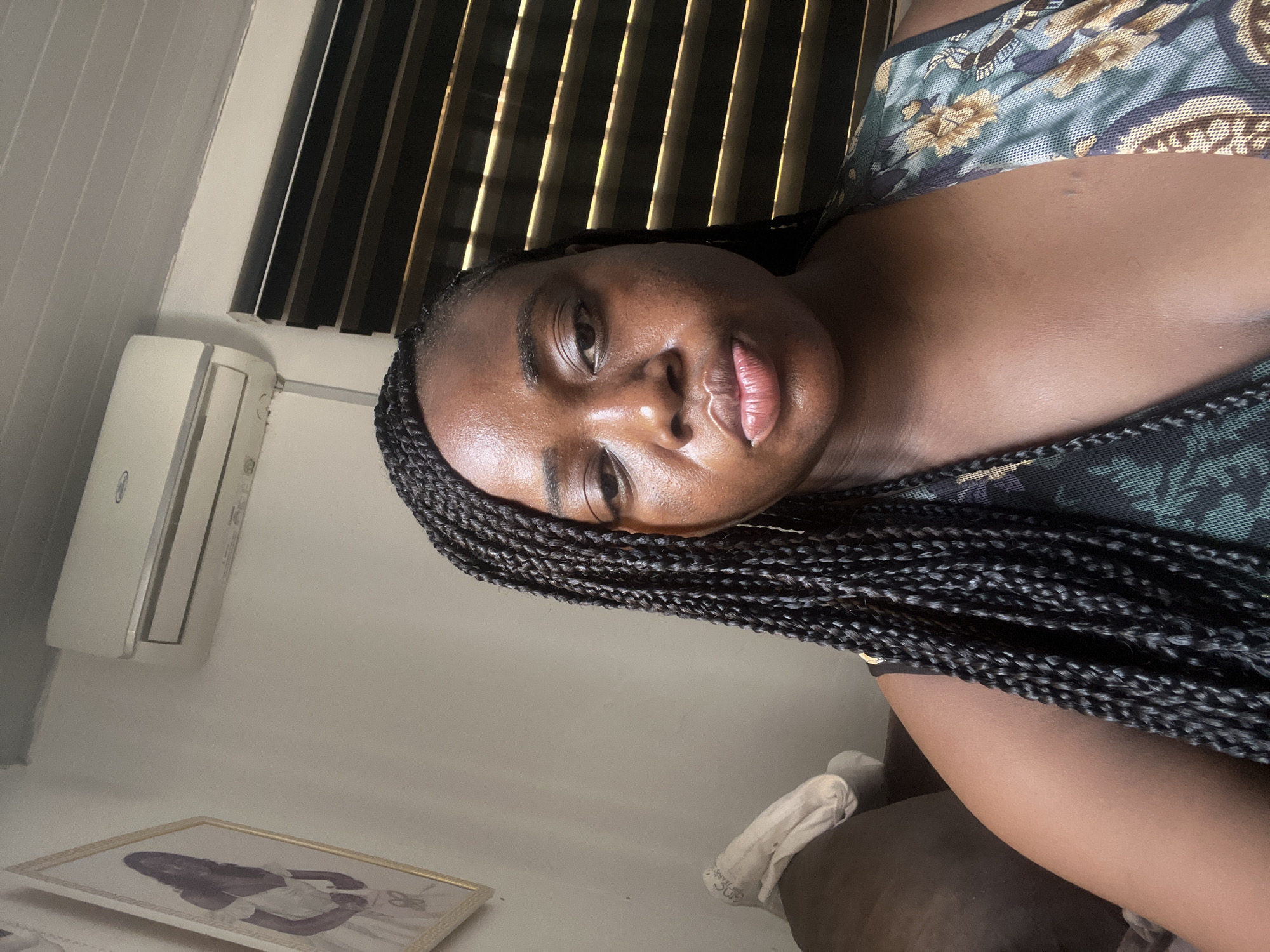 A close-up, tilted portrait features a dark-skinned woman with a relaxed, neutral expression, possibly with a slight pout. Her face appears somewhat shiny, and she has long, dark box braids styled over her shoulder. She is wearing a patterned top with floral or intricate designs in shades of green, blue, and gold.
The setting is indoors. In the background, strong sunlight filters through horizontal blinds, casting distinct stripes of light and shadow. To the woman's upper left, a white wall-mounted air conditioning unit is visible against a light-colored wall. Towards the lower left of the frame, there is an ornate golden picture frame resting on a surface, containing what appears to be a black-and-white or sepia-toned image of a woman in a light-colored dress. The woman in the main image appears to be reclining on a dark surface, possibly a bed or sofa, with a piece of white fabric, possibly a sock, visible in the lower right.
The image is tilted counter-clockwise, suggesting the photo was taken while the subject was lying down or reclining. The visible sunlight indicates it is daytime. The overall mood is calm and relaxed. No discernible text is visible.