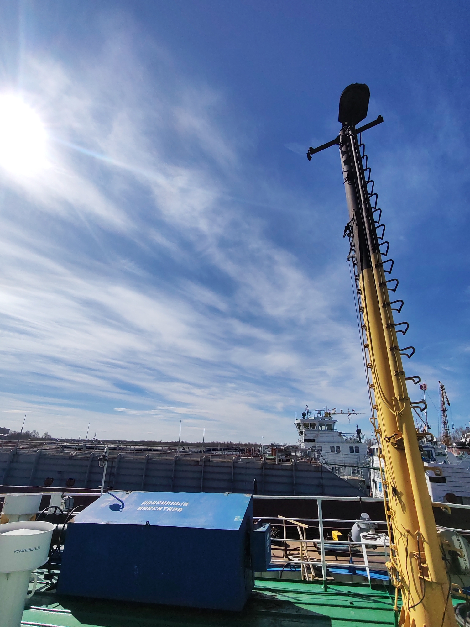 The image captures a bright, sunny daytime scene at a port or shipyard in Rybinsk, Russia. The sky is a vivid blue with wispy white cirrus clouds stretching across it. A brilliant sun, located in the upper left, casts strong light and creates a prominent lens flare.
In the foreground, the green deck of a vessel is visible, with a sturdy blue box resting on it. Written in white Cyrillic letters on the blue box is "АВАРИЙНЫЙ ИНВЕНТАРЬ," which translates to "Emergency Inventory" or "Emergency Equipment." To the left of the blue box, a white cylindrical object with "РУМПЕЛЬНОЕ" (likely related to "Rudder" or "Steering Gear") written on it can be seen. A white safety railing spans across the middle distance.
Dominating the right side of the frame is a tall, thick yellow mast or boom, equipped with a series of black rungs or ladder steps running along its length. At its apex, a dark, possibly metallic, object resembling a sensor or light fixture is mounted.
In the mid-ground, behind the foreground vessel's deck, stands a white and grey ship, possibly a tugboat or service vessel, featuring a multi-deck superstructure with a bridge area and various antennae or radar equipment. Further back, a large, dark grey hull, likely a barge or cargo ship, is moored alongside a quay or dock. In the distant background, industrial structures and cranes are faintly visible under the bright sky. The overall impression is one of maritime activity under clear, pleasant weather conditions.