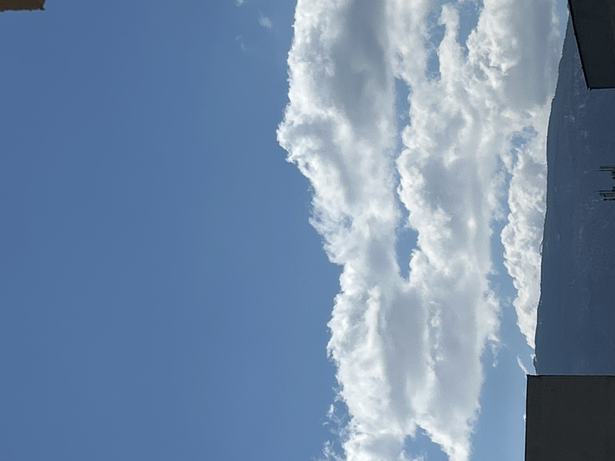 The image shows a vertical view of a blue sky with fluffy white clouds. A dark gray building is partially visible on the right side of the frame. The image could be taken in an urban area. Sofia, Bulgaria cannot be determined based on this image.