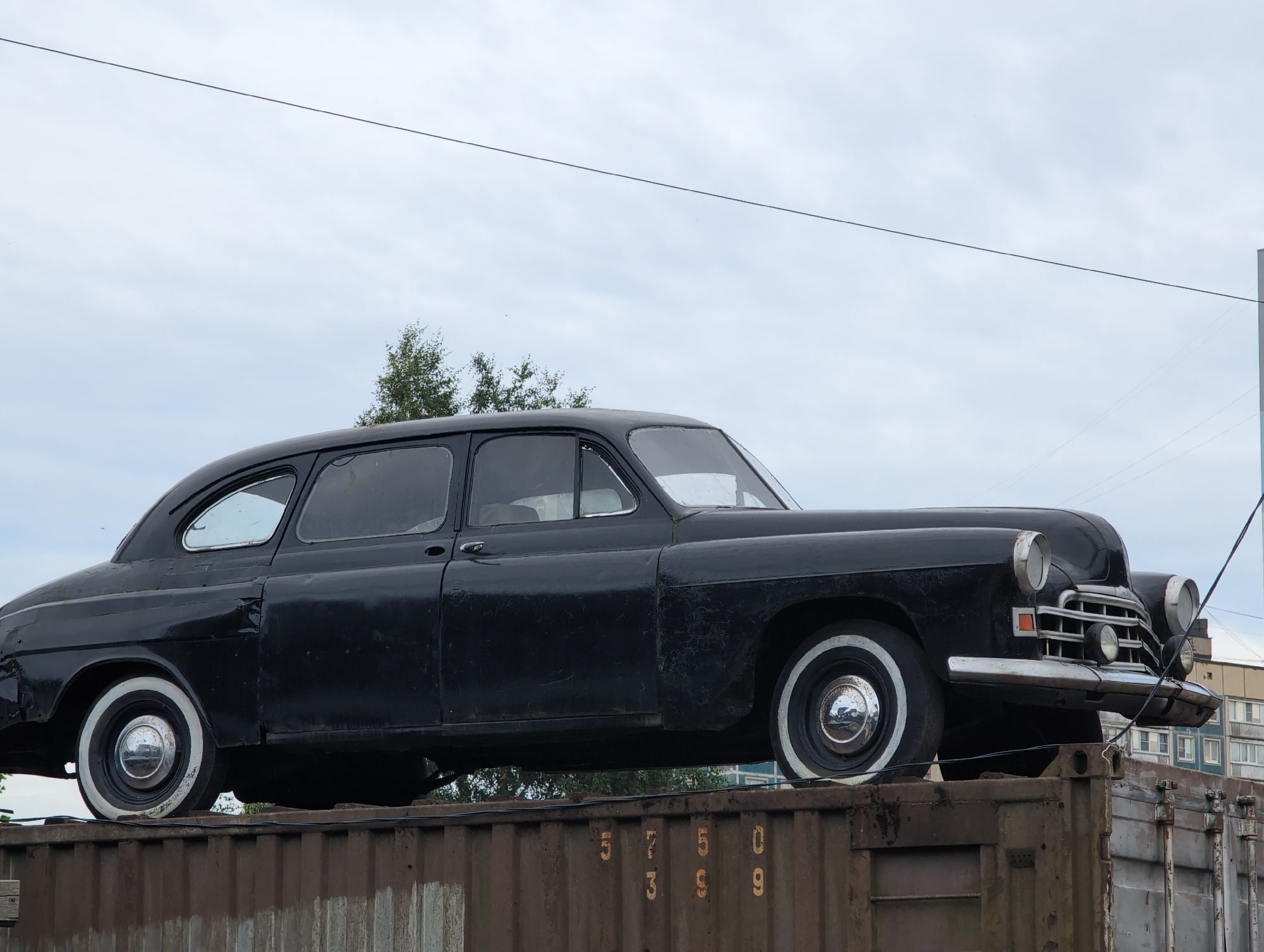 A black vintage automobile with whitewall tires is positioned atop a weathered, rusty shipping container. The car, viewed from its side, features chrome accents and rounded body lines characteristic of mid-20th-century design. Behind the car, a building with a blue facade and multiple windows is partially visible, suggesting an urban or industrial setting. The sky above is overcast with scattered clouds. The shipping container displays faded yellow stenciled numbers, reading "573 59 09". The scene appears to be located in Novoe Devyatkino, Russia, with overhead power lines visible against the sky.