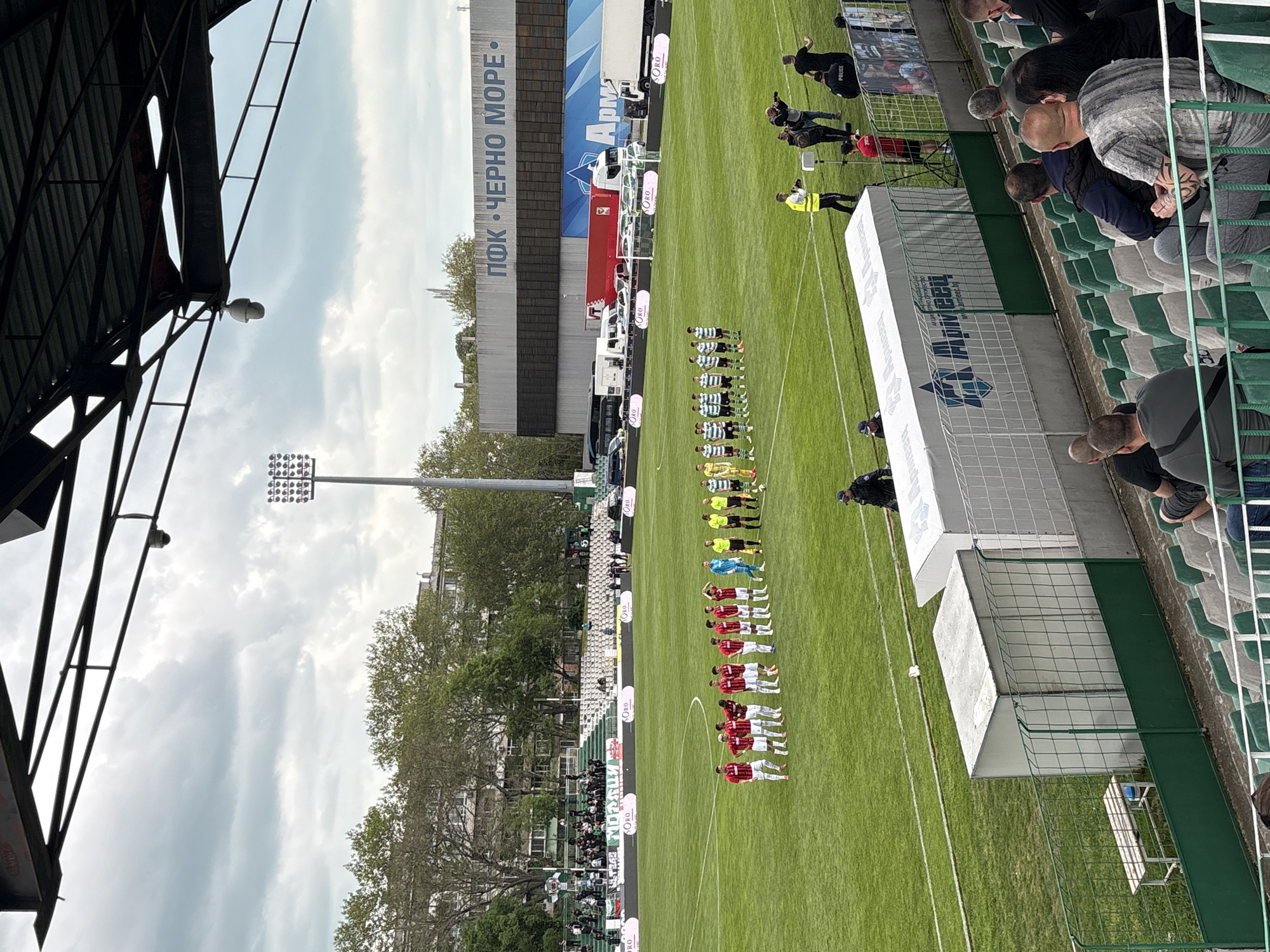 Here's a description of the image:
This image shows a view of a soccer stadium in Varna, Bulgaria. Two soccer teams in their respective red and green uniforms line up on a grass field. Spectators are seated in the stands on the sidelines. The stadium features advertising banners and a large sign reading "ПФК ЧЕРНО МОРЕ," which translates to "PFC Cherno More" (a local soccer team). A tall lighting tower stands near the field. The sky above is overcast. The image is taken at an angle, showing parts of the stadium's roof.