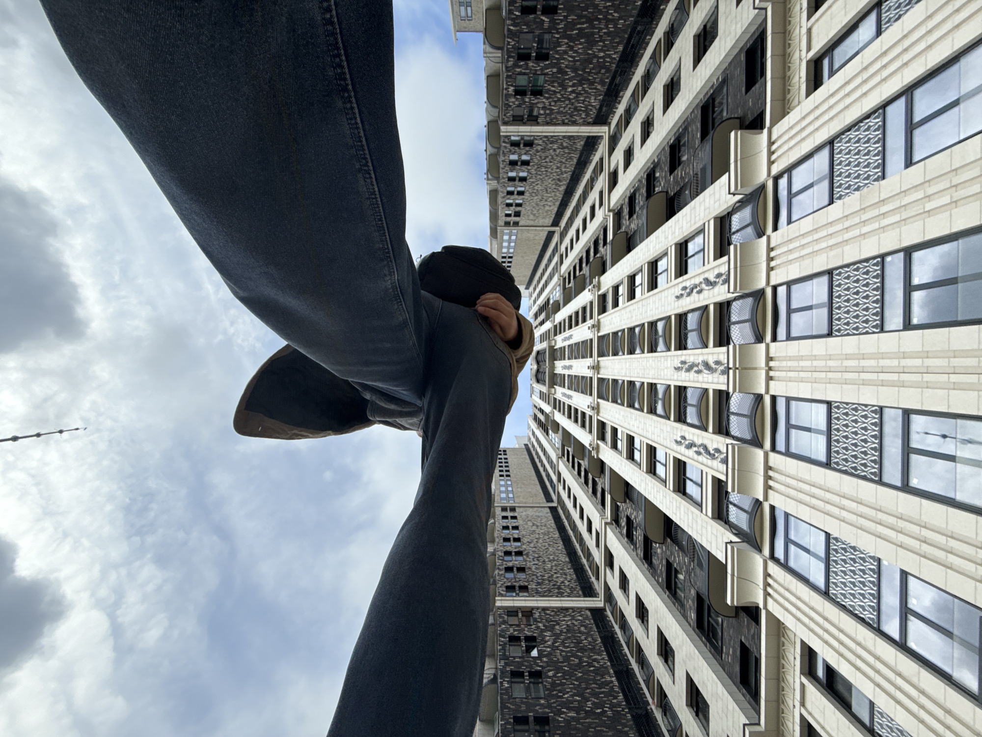 A low-angle shot captures a person wearing dark jeans and boots, positioned seemingly at the edge of a tall building. Above the person, a building with a light-colored facade featuring rows of windows with dark frames is visible. A darker, brick-like structure can be seen adjacent to the lighter building. The sky is partly cloudy. The location cannot be determined from the image.