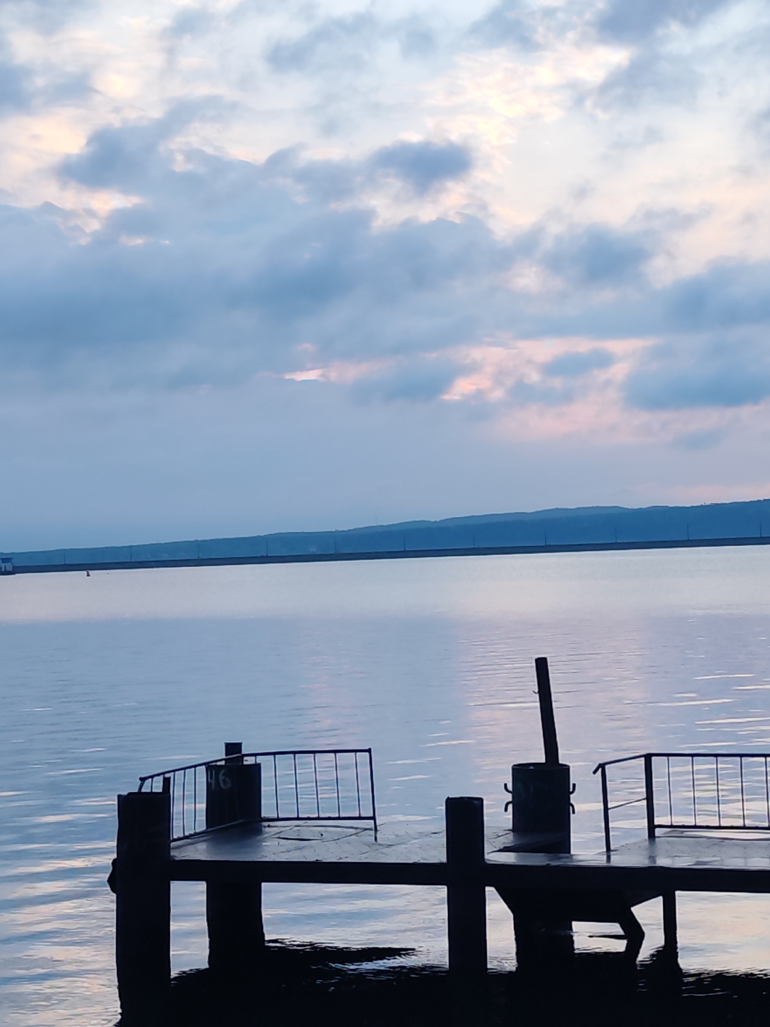 The image shows a tranquil scene on the Kama Reservoir in the city of Chaikovsky, Russia. A weathered pier juts out into the calm, reflective water. The pier is constructed of dark wood pilings and has metal railings along its sides. A metal can-shaped object with metal hooks attached to it stands on the pier. A number "46" is visible on the pier. In the background, the reservoir stretches to the horizon where a long dam or levee runs along the edge of the reservoir. The sky is filled with gray and blue clouds, with hints of pink indicating either dawn or dusk. The light is soft and diffused, creating a peaceful atmosphere. The scene is devoid of people.